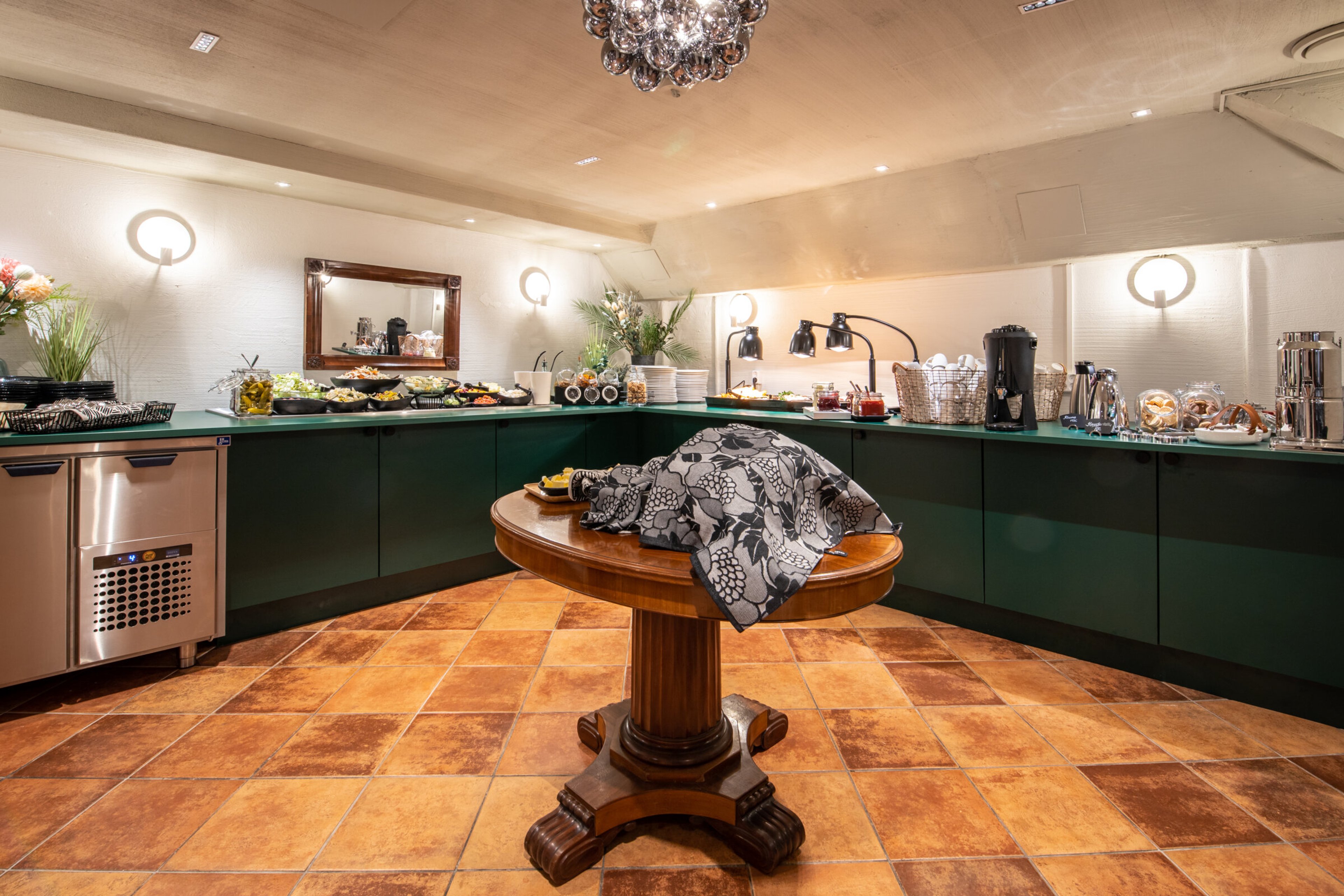 Tarjoilupöytä ravintolan buffet-tilassa, jossa vihreät työtasot, koristeellinen kattokruunu ja terrakottalaattalattia. Keskellä puinen pöytä.