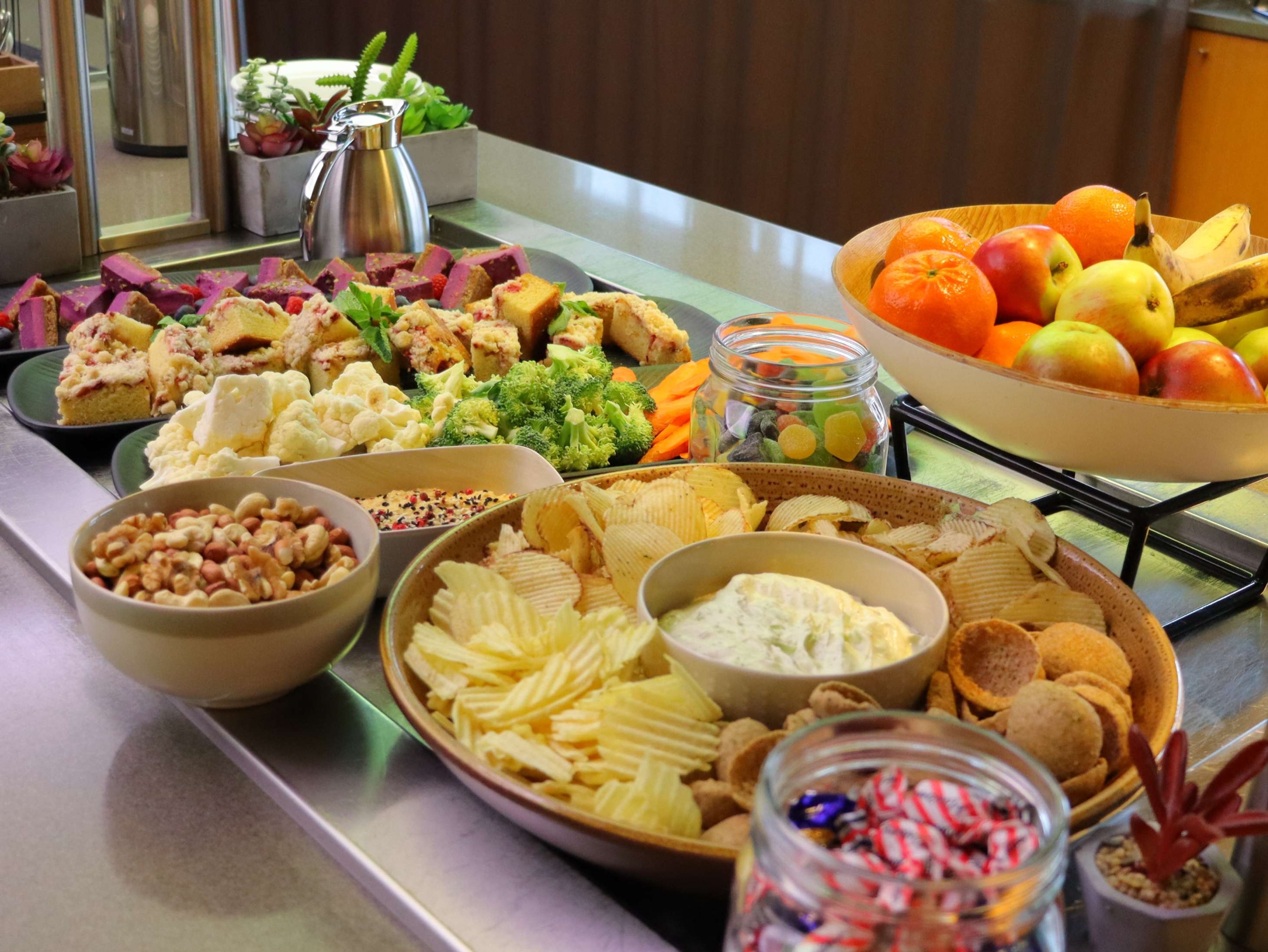 Monipuolinen tarjoilupöytä, jossa leivonnaisia, sipsejä, dippiä, pähkinöitä, vihanneksia ja hedelmäkulho täynnä omenoita, banaaneja ja appelsiineja.