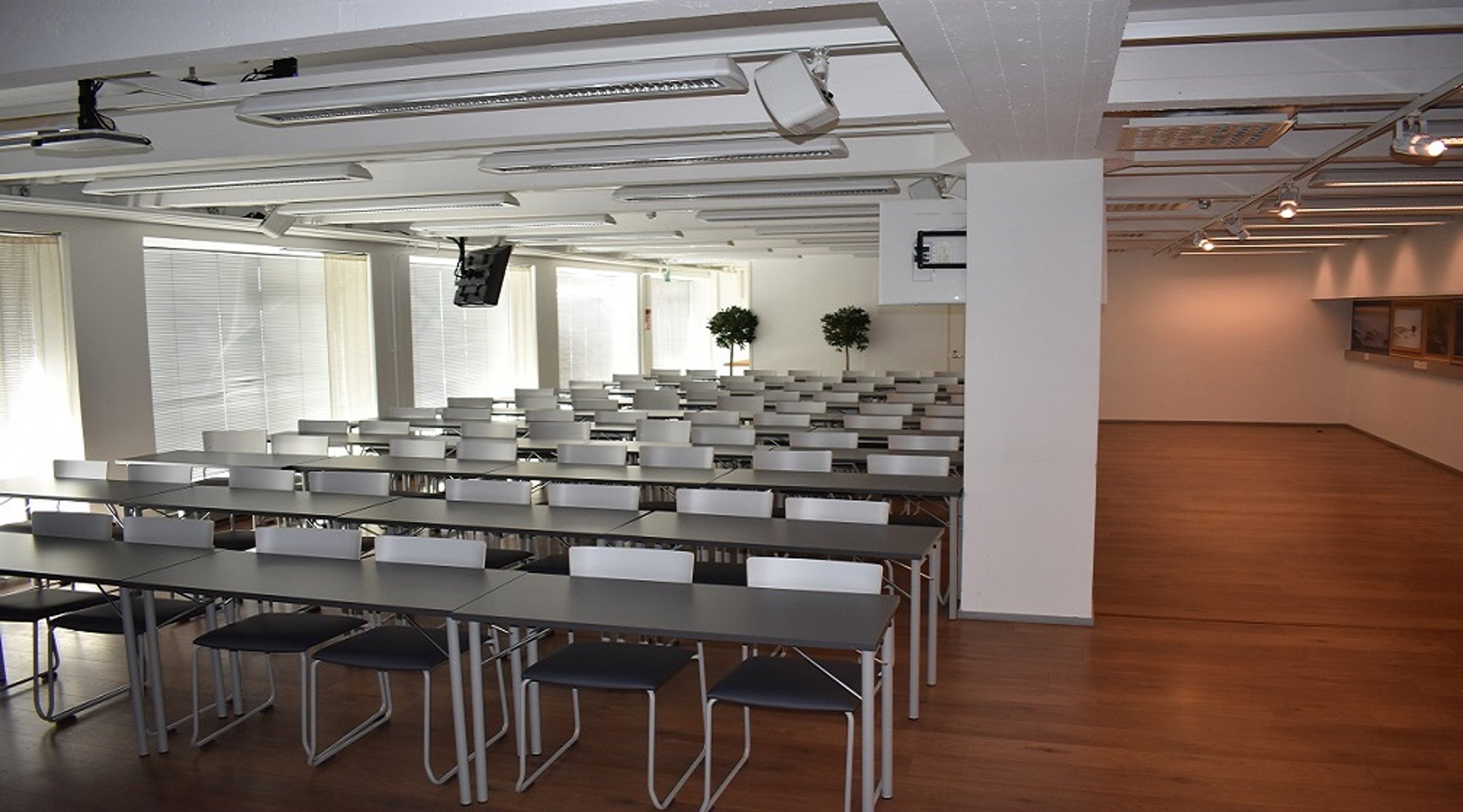 Valoisa koulutustila, jossa harmaat pöydät ja mustat tuolit riveittäin. Puulattia ja valkoinen tila jatkuu pilarin toisella puolella.