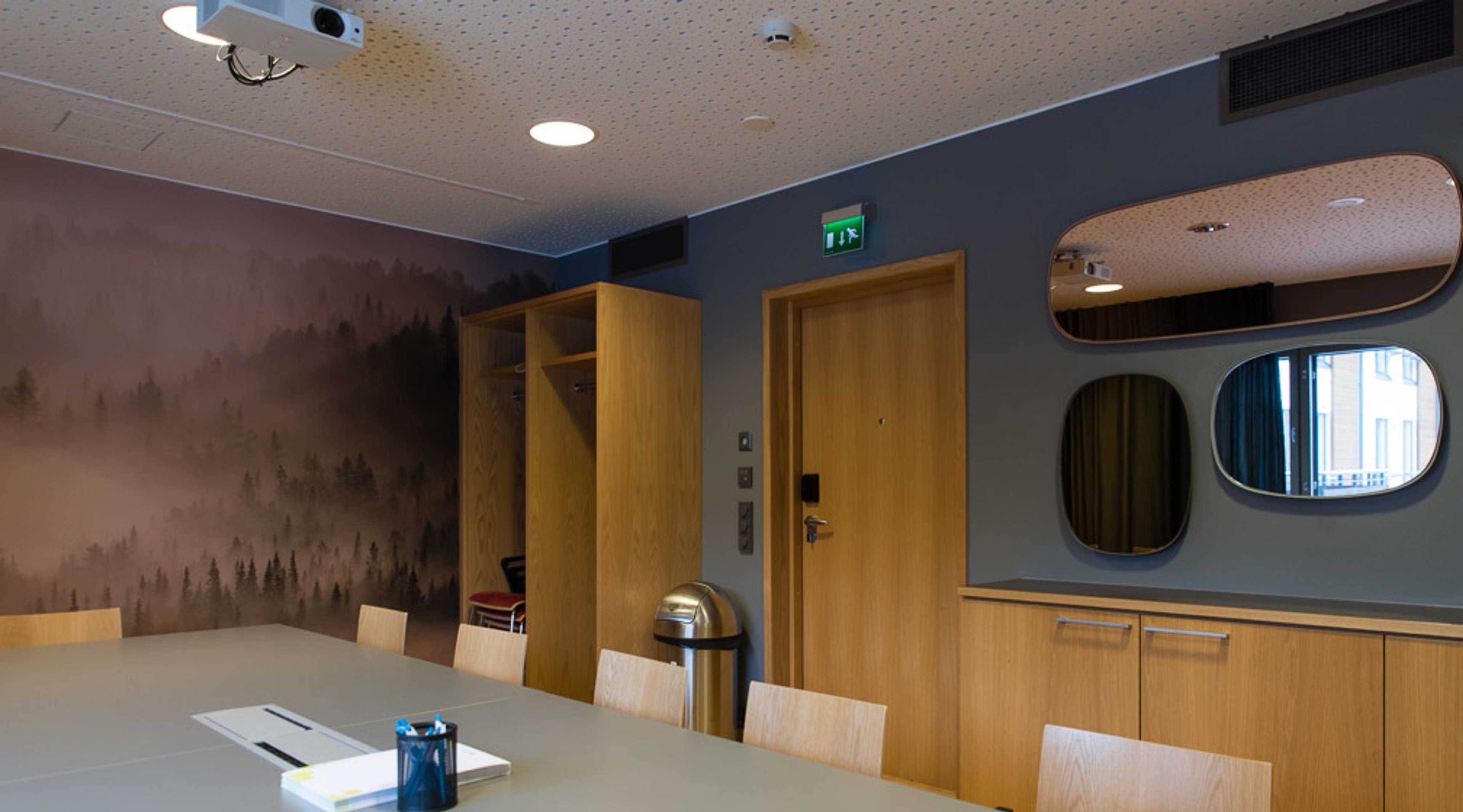 Moderni neuvotteluhuone puisella pöydällä, tuoleilla ja kalusteilla. Seinällä metsäsumumaalaus, katossa projektori ja tasaisella lattialla.