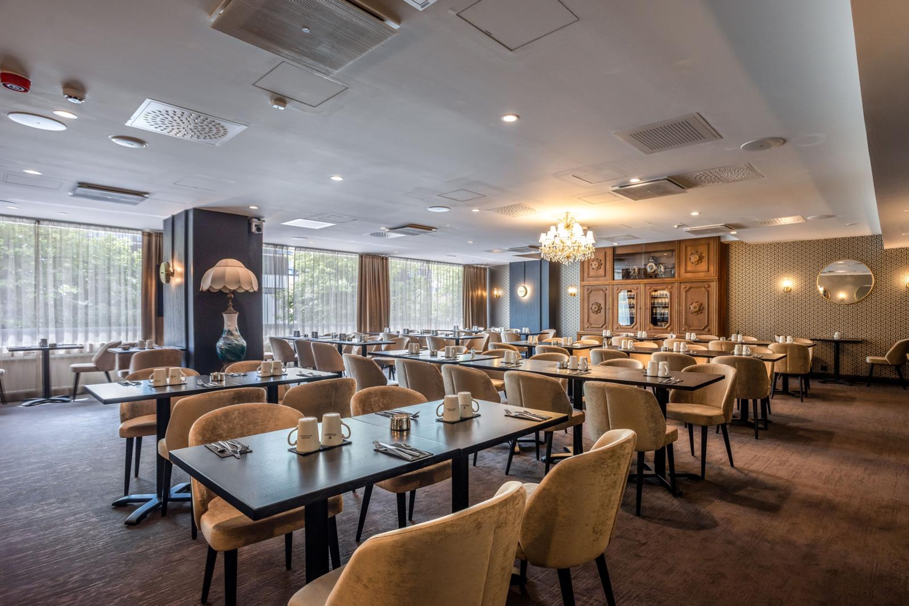 Elegantti ravintolan ruokasali, jossa on pehmeät beiget tuolit, mustat pöydät ja kattokruunu. Tilassa on laadukas sisustus, puinen seinäkaappi ja suuret ikkunat.