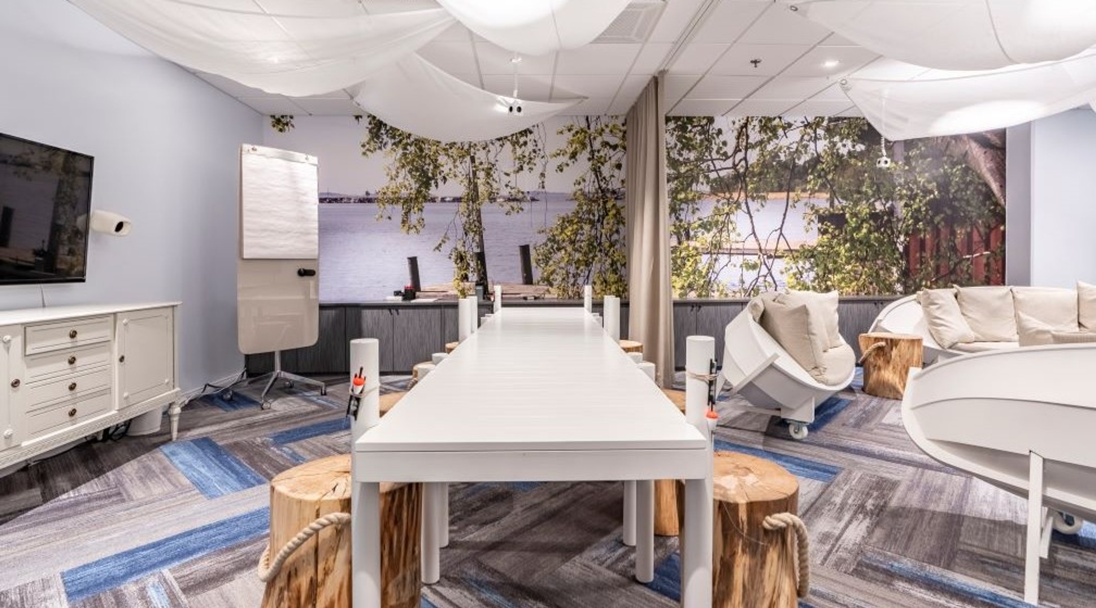 Moderni neuvotteluhuone, jossa pitkä valkoinen pöytä, puisia istuimia ja seinällä luontomaisematapetti. Kattoa koristavat valkoiset kankaat ja tilassa on taulutelineitä.