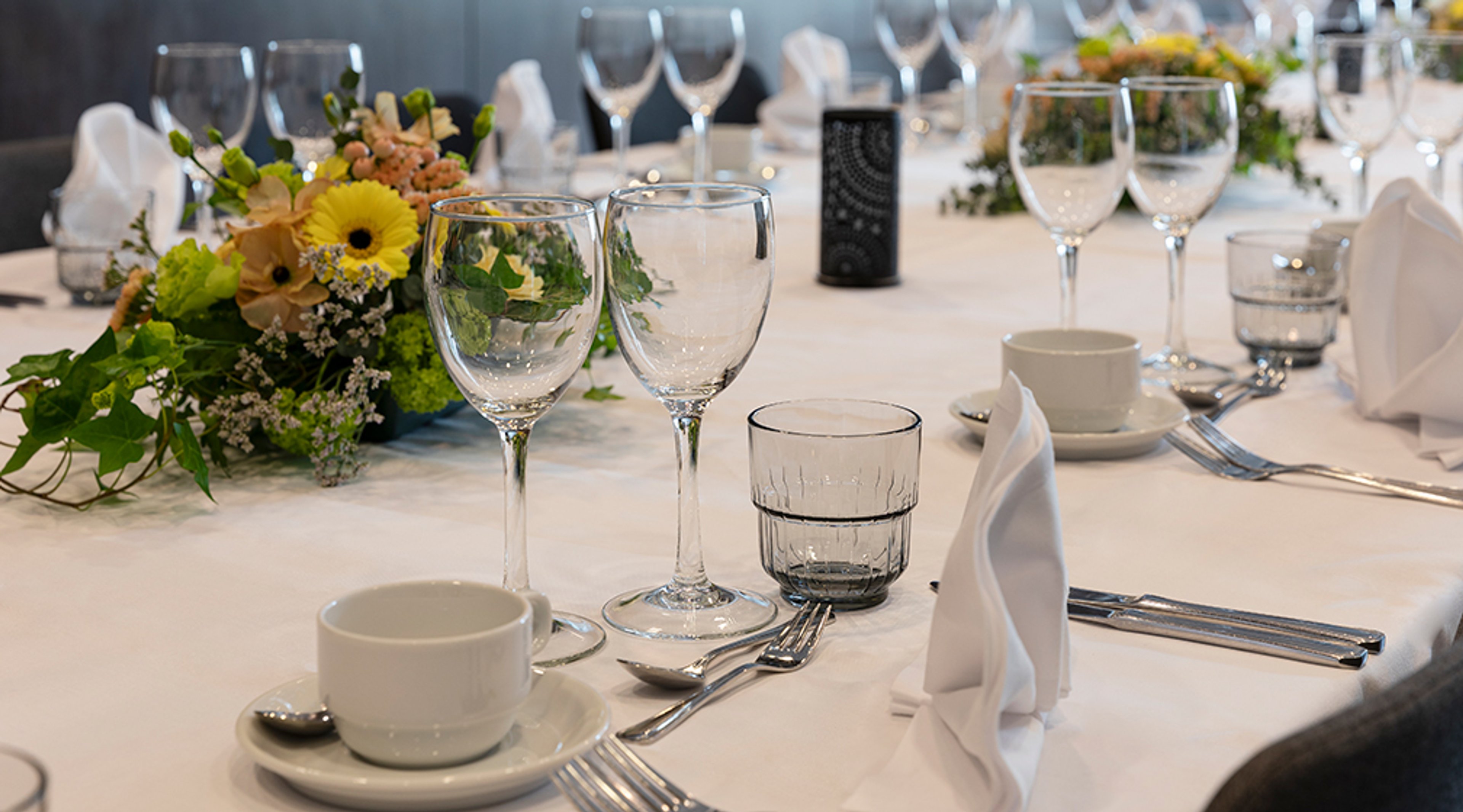 Juhlallisesti katettu pöytä valkoisella liinalla, kauniiden kukkakimppujen, viinilasien, kahvikuppien ja aterimien kera.