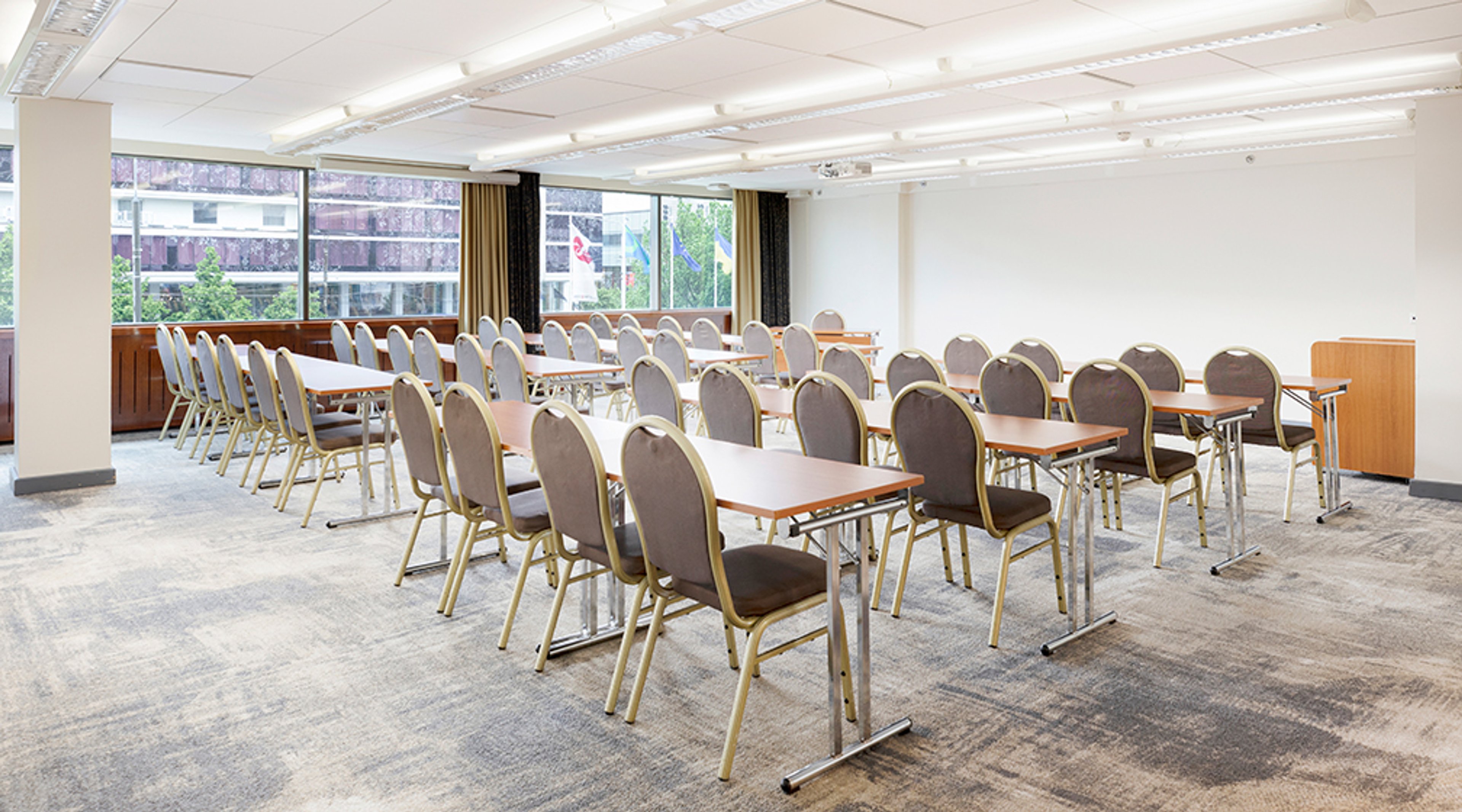 Conference room with rows of tables and chairs on hard flooring. Room has large windows providing natural light.