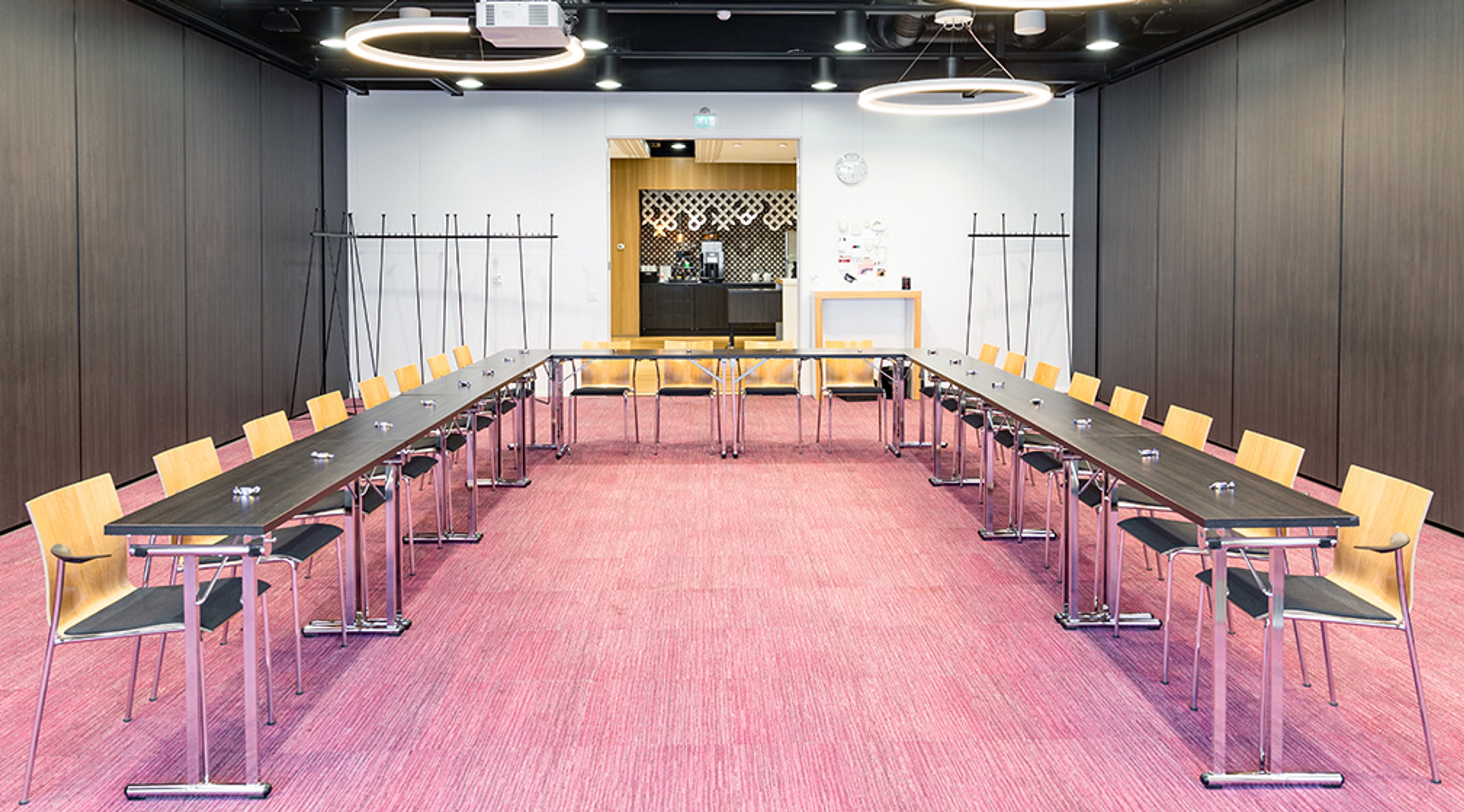 Moderni kokoushuone, jossa U-muotoinen pöytäjärjestys, puiset tuolit ja pinkin värinen lattia. Laajat kulkuväylät ja tasainen lattiapinta.