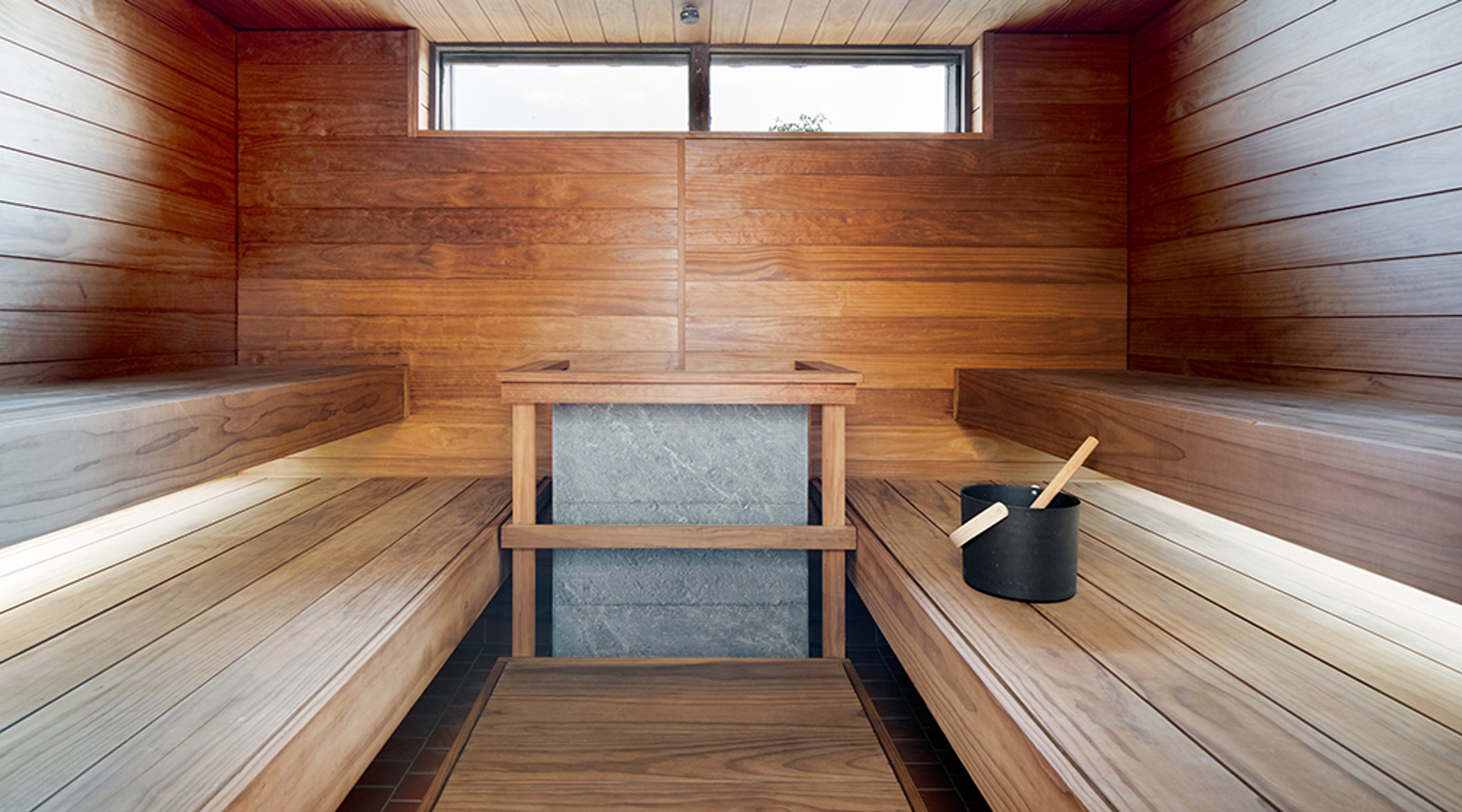 Moderni sauna puupaneeliseinillä, istumatasot molemmilla sivuilla, keskellä kivikiuas ja ylhäällä ikkunarivi.