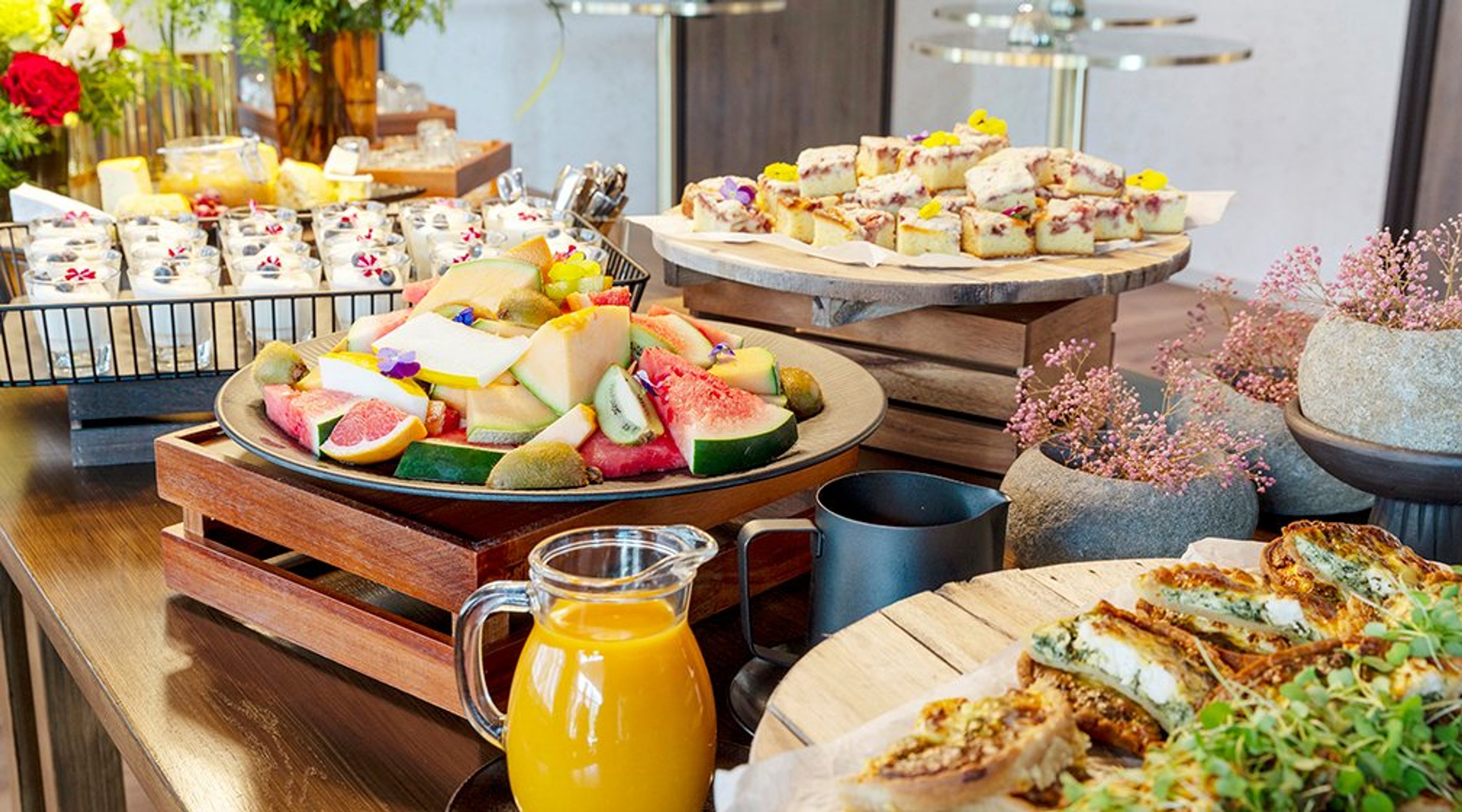 Kattava aamiaispöytä, jossa leikattuja hedelmiä, leivoksia, pientä suolaista ja kannullinen mehua puisella tarjoilupöydällä.