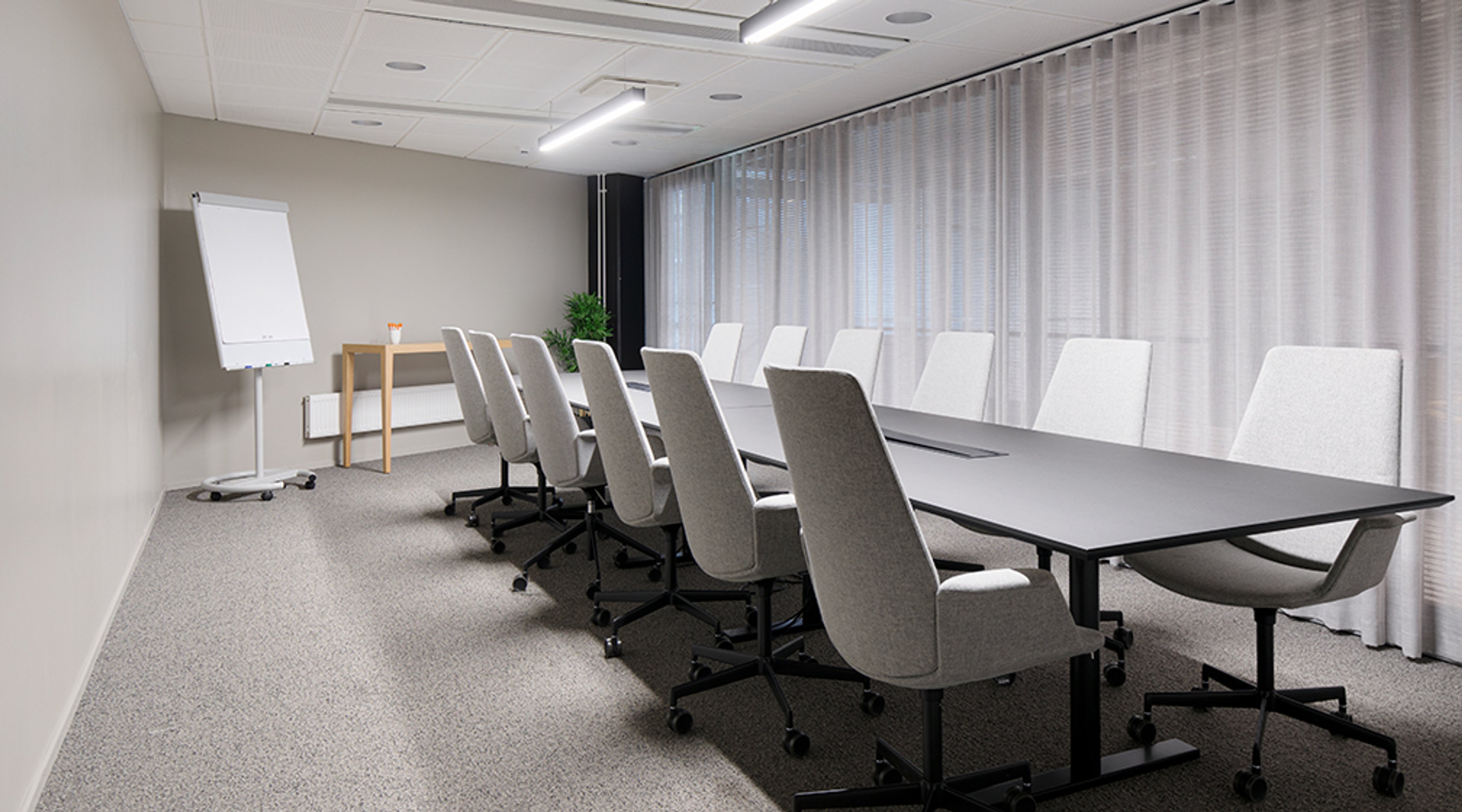 Modern meeting room with long black table and light office chairs, flip chart in corner and light curtains covering large windows.