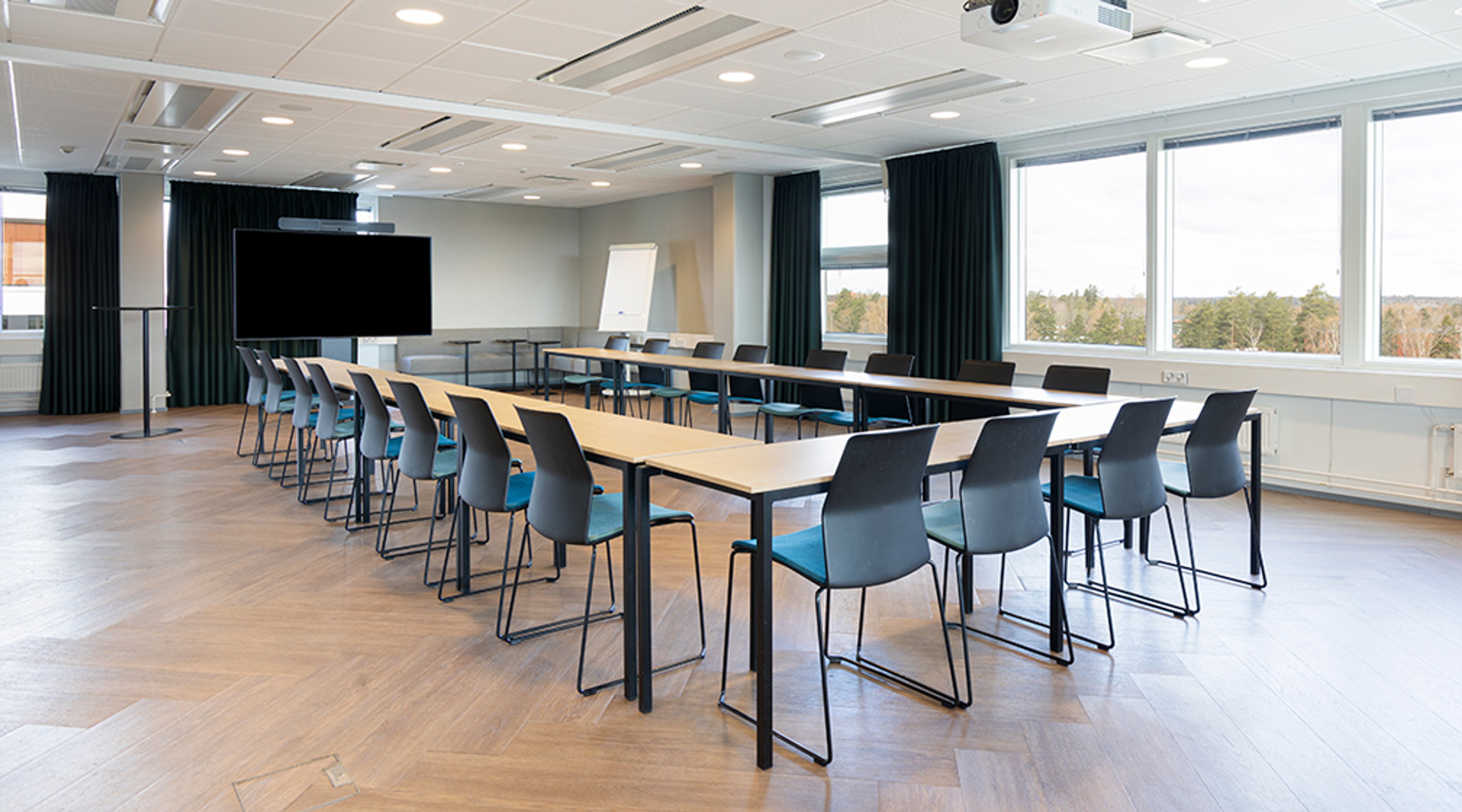 Meeting room with a long table and blue chairs, large screen and whiteboard on the wall, and large windows with forest view. Hardwood floor and good lighting.
