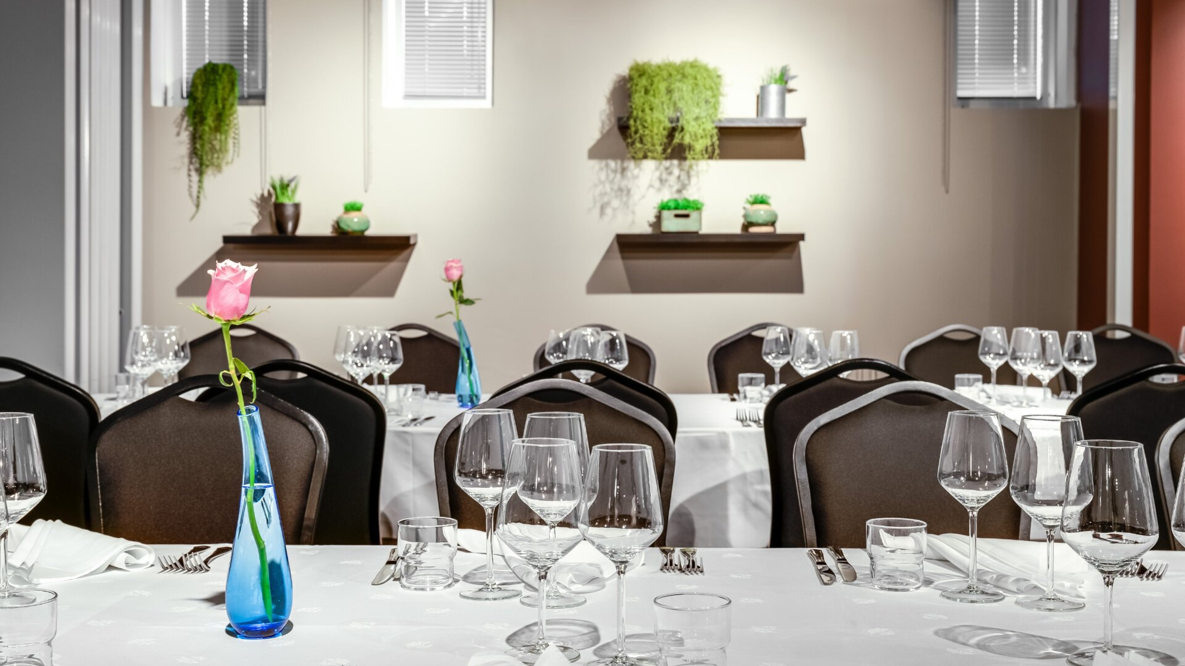 Ravintolan ruokailutila valkoisilla pöytäliinoilla, viinilaseilla ja sinisellä maljakolla, jossa vaaleanpunainen ruusu. Seinällä viherkasveja hyllyillä.