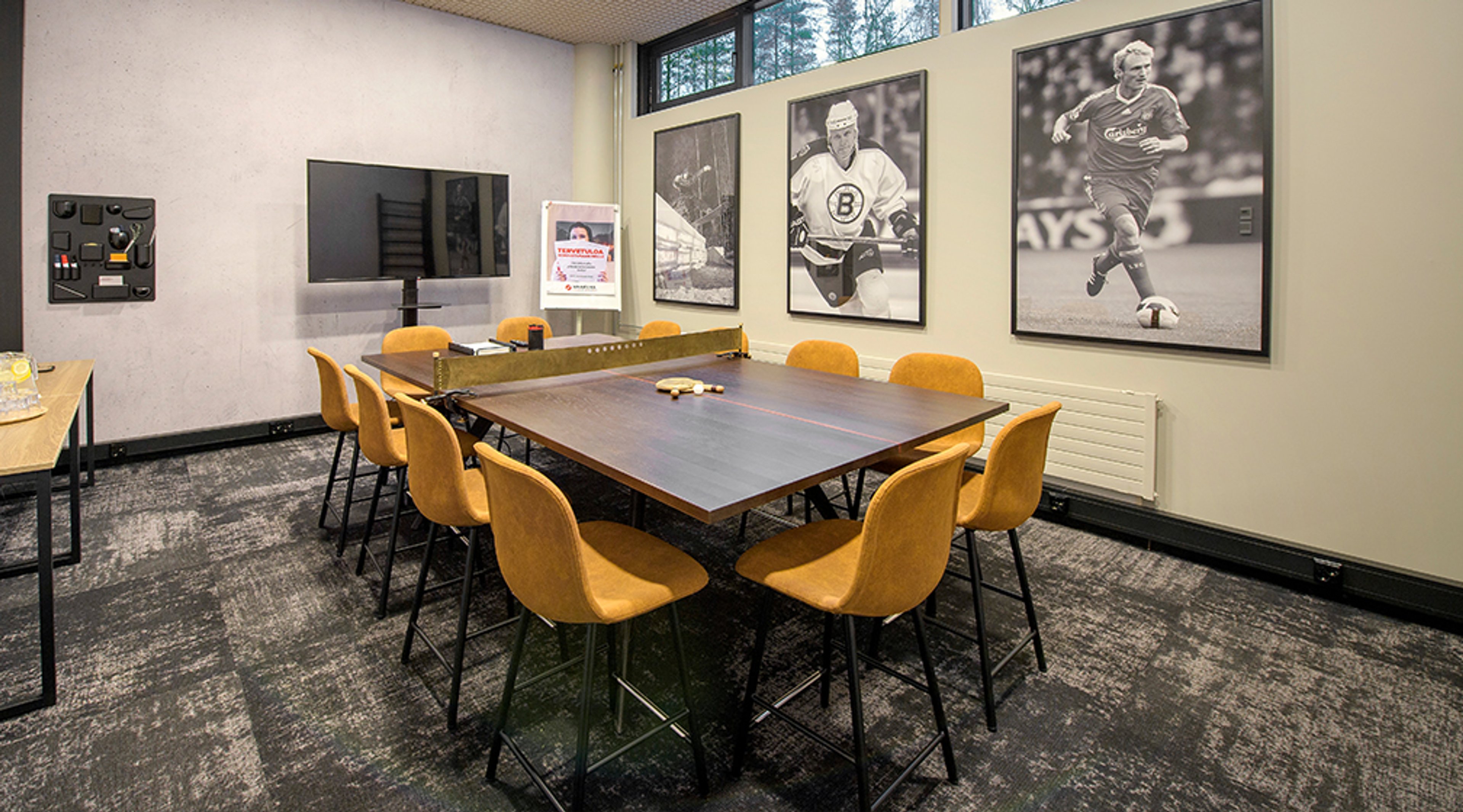 Neuvotteluhuone, jossa puinen pöytä ja keltaiset tuolit. Seinillä urheiluaiheiset mustavalkokuvat ja tilassa on näyttö esityksiä varten.