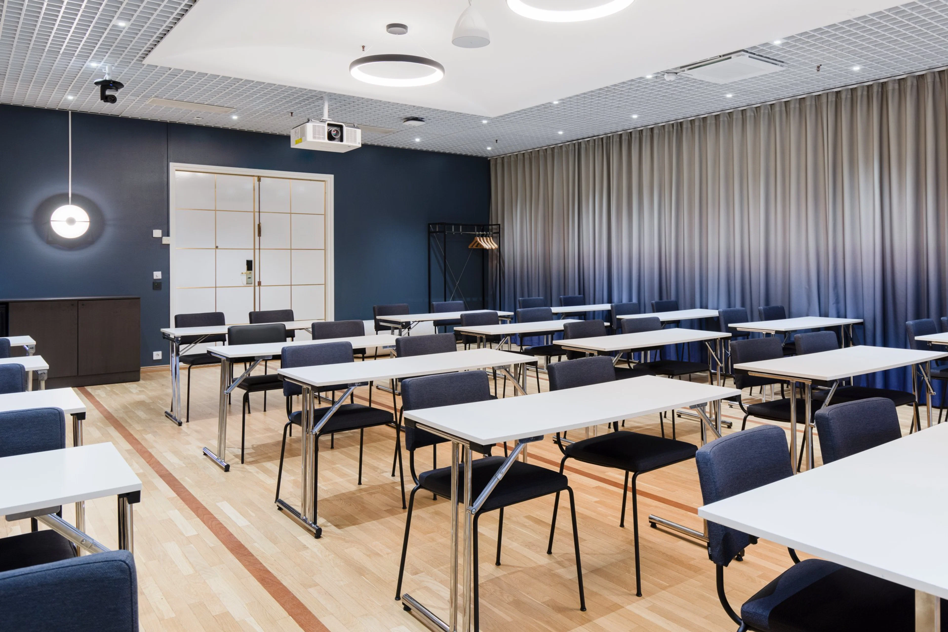 Moderni kokoustila sinisillä seinillä, puulattialla ja valkoisilla pöydillä. Tilassa projektori, verhot ja tasainen kulkureitti.
