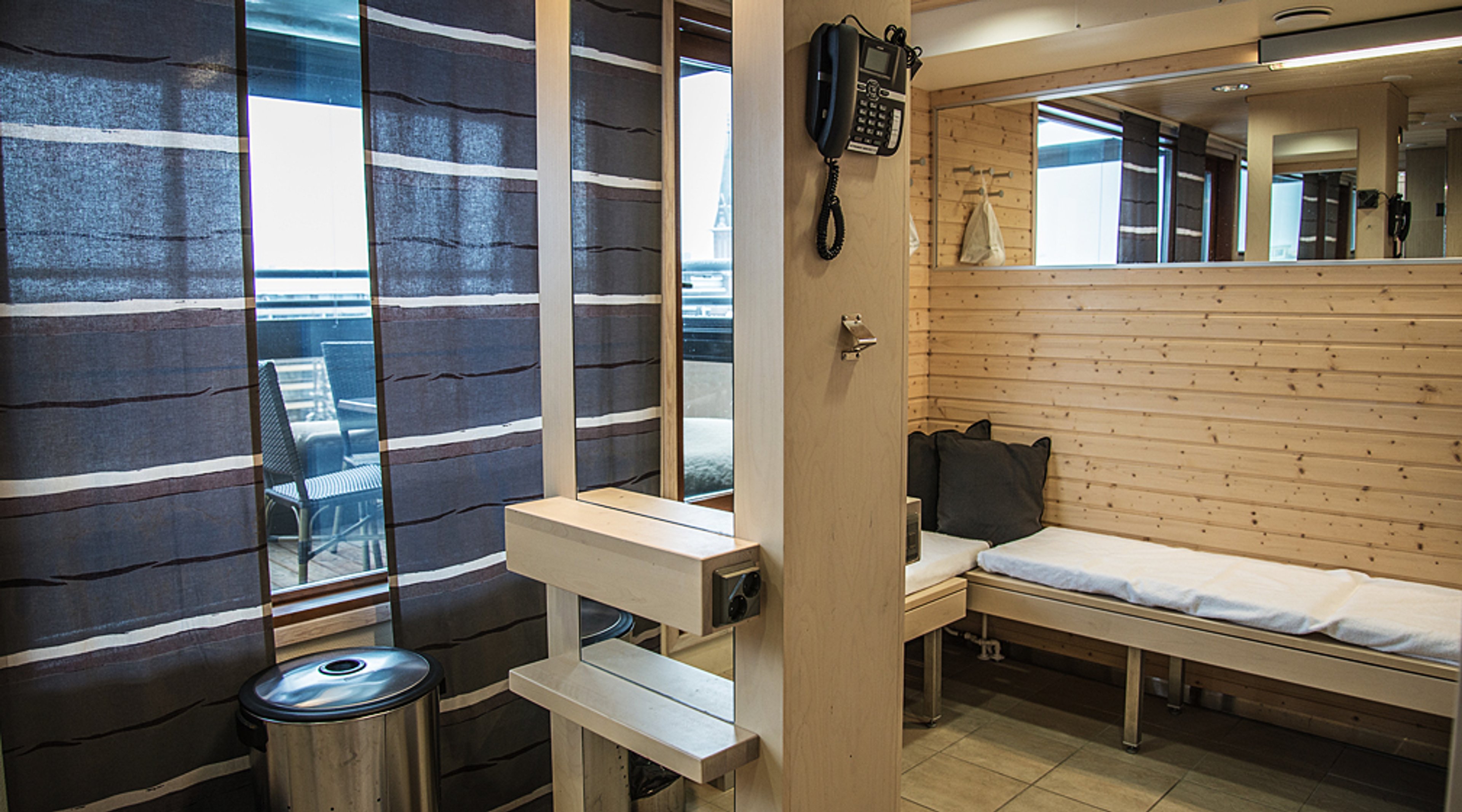 Sauna- tai hoitohuone puupaneeliseinillä, siniraitaisilla verhoilla ja pitkällä penkillä. Ikkunasta näkyy ulkotila.