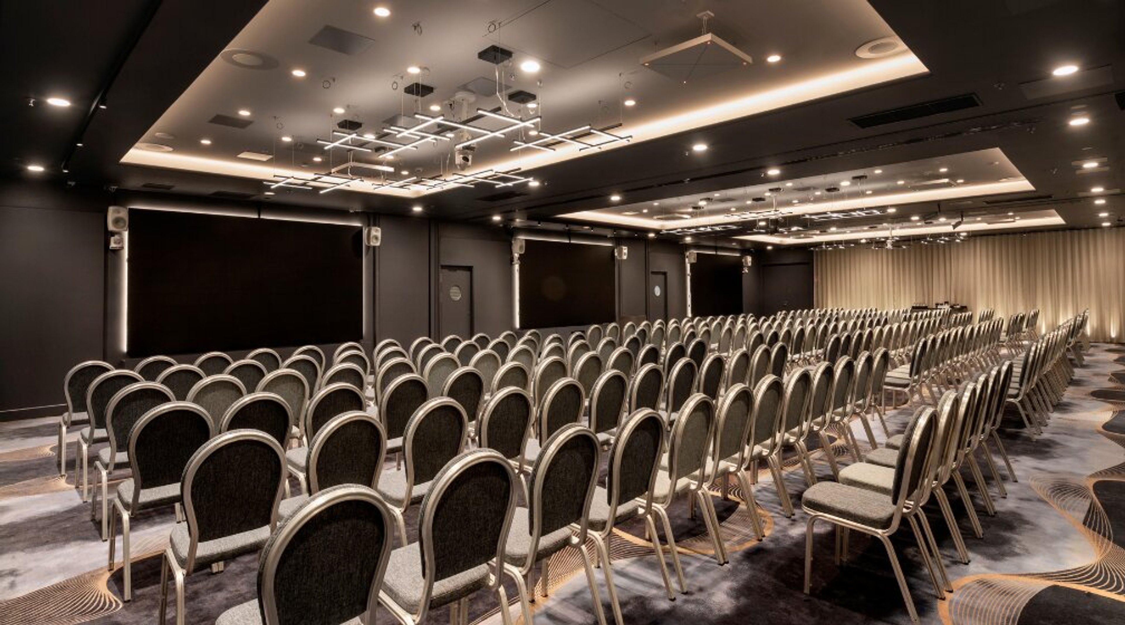 Moderni auditorio riveittäin järjestetyillä tuoleilla, tummilla seinillä ja tyylikkäällä valaistuksella katossa.
