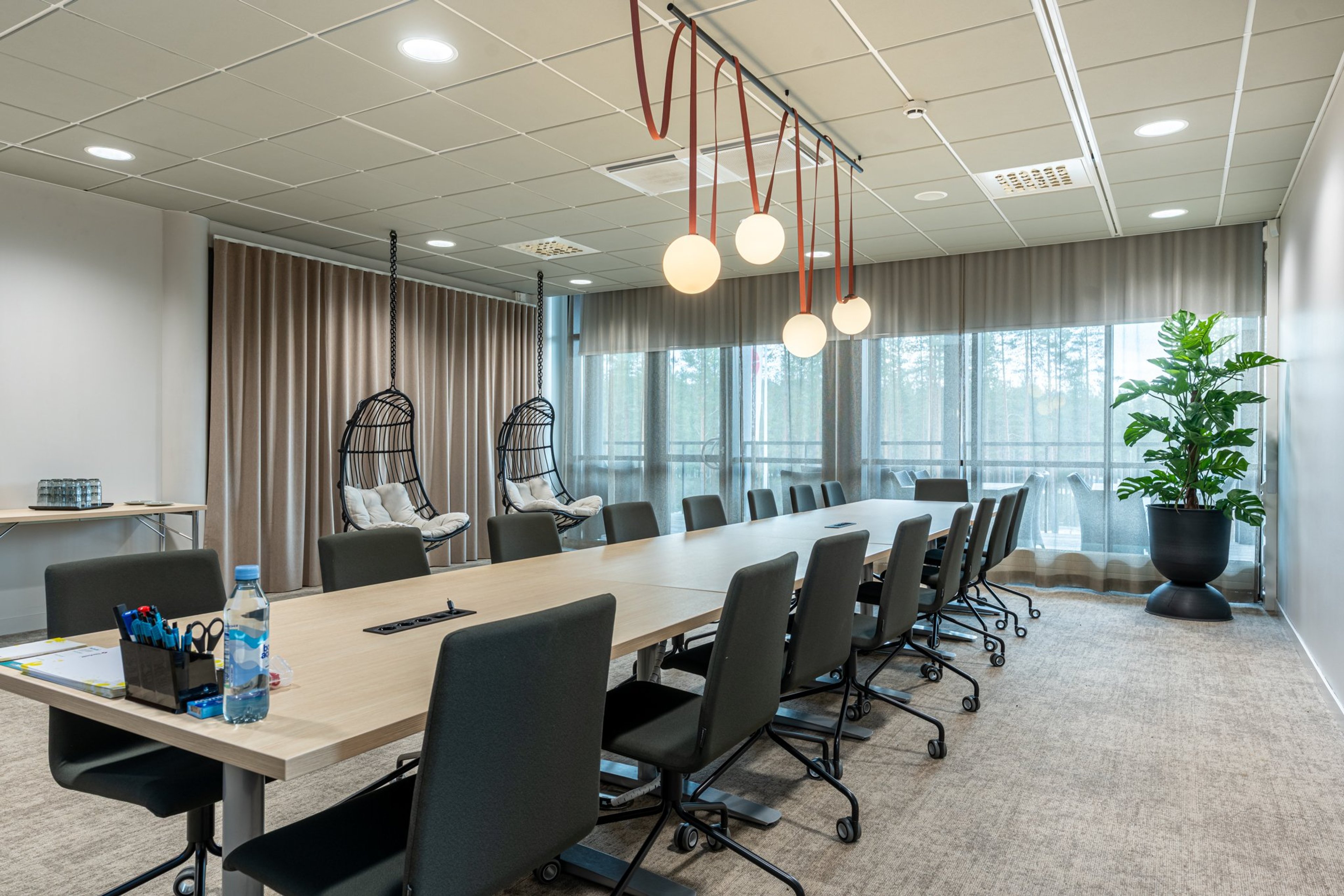 Moderni neuvotteluhuone, jossa pitkä puinen pöytä, pyörälliset tuolit, riippuvat nojatuolit ja pyöreät riippuvalaisimet. Suurista ikkunoista näkyy metsämaisema.