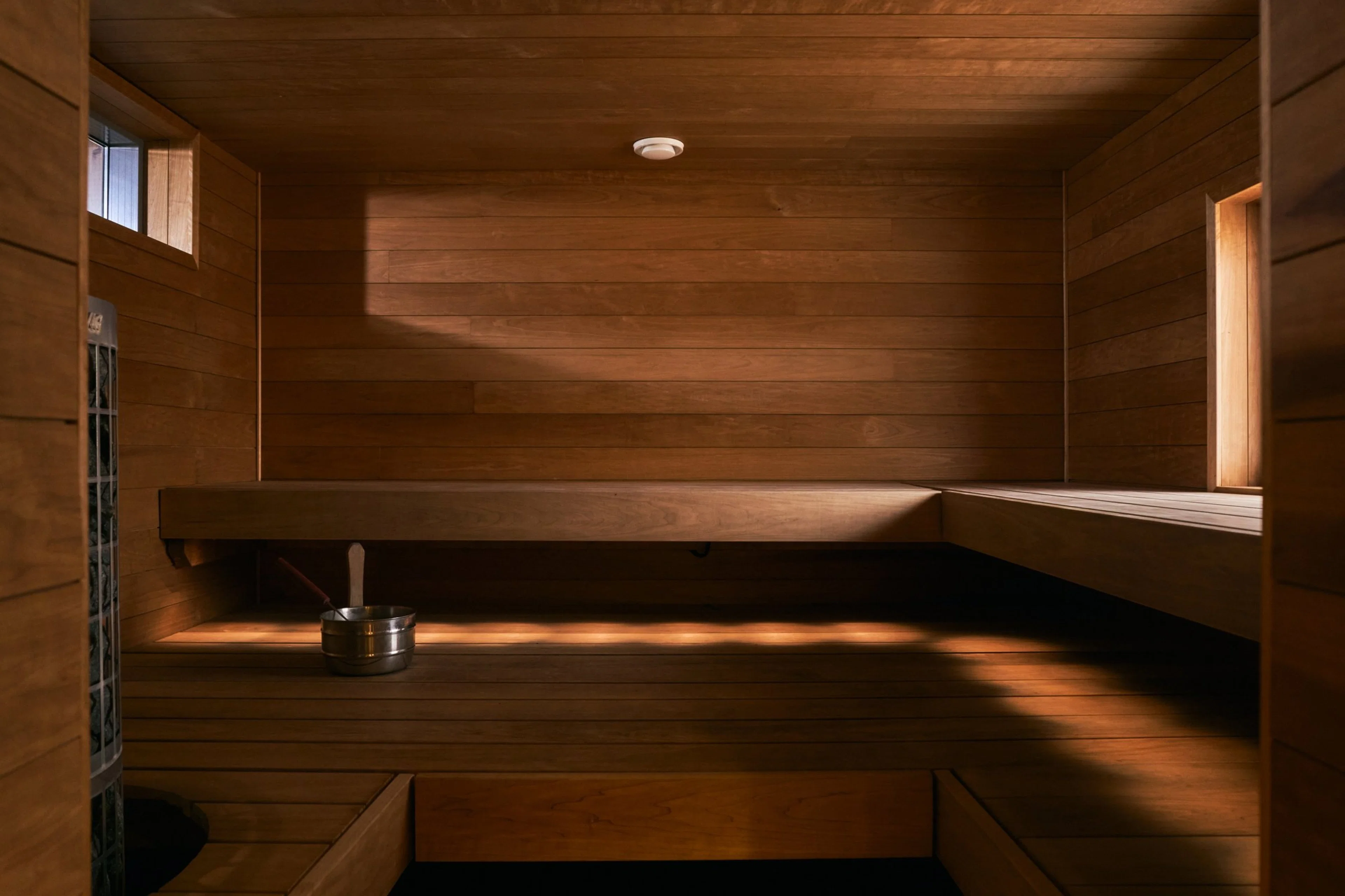 Puupaneloitu sauna L-muotoisilla istuintasoilla, seinällä kiuas, lattia- ja kattotasot puuta, valaistus luo lämpimiä varjoja.