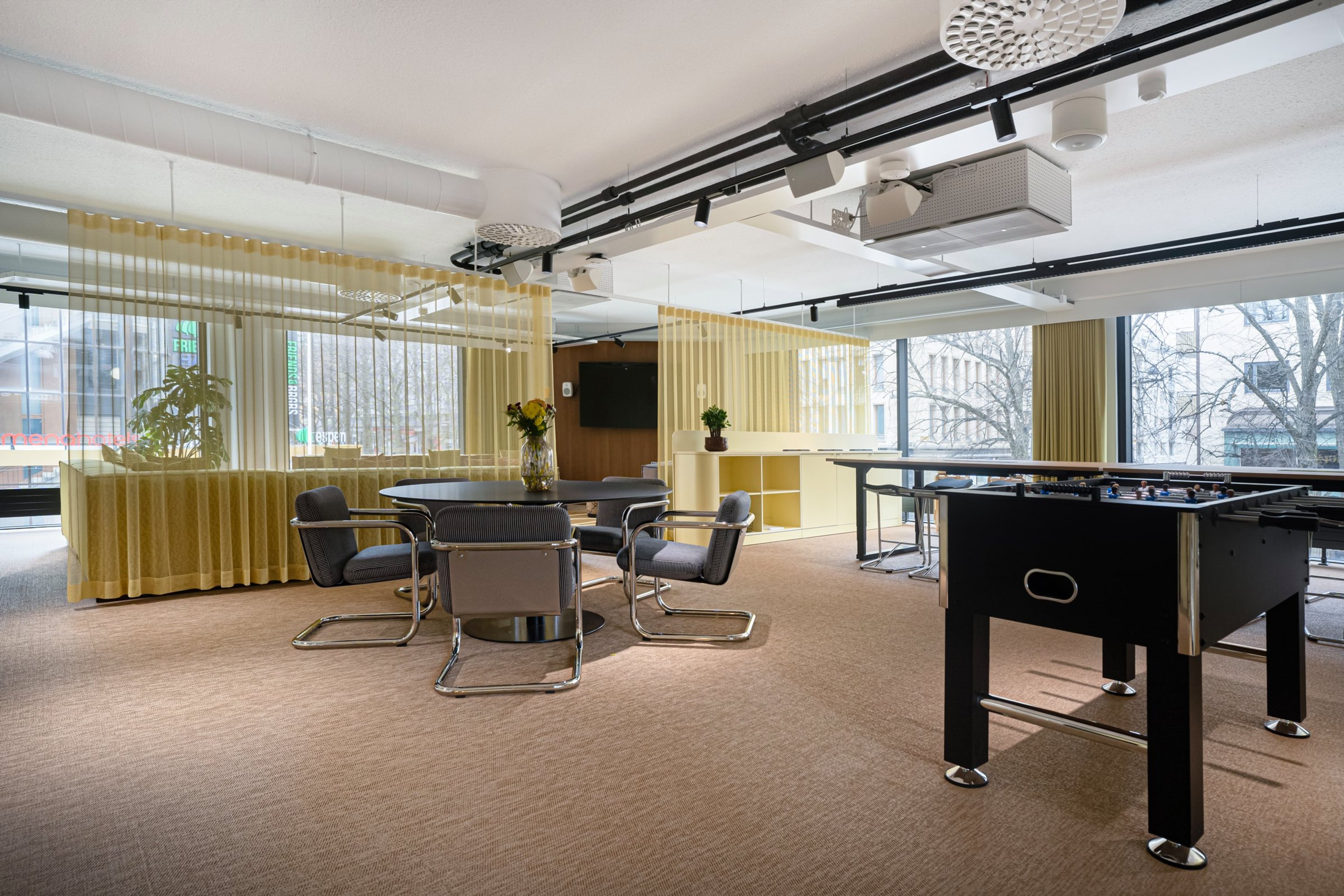 Moderni taukotila, jossa pyöreä pöytä ja tuoleja, pöytäfutispeli ja keltaiset verhot. Tasainen kokolattiamatto ja isot ikkunat.