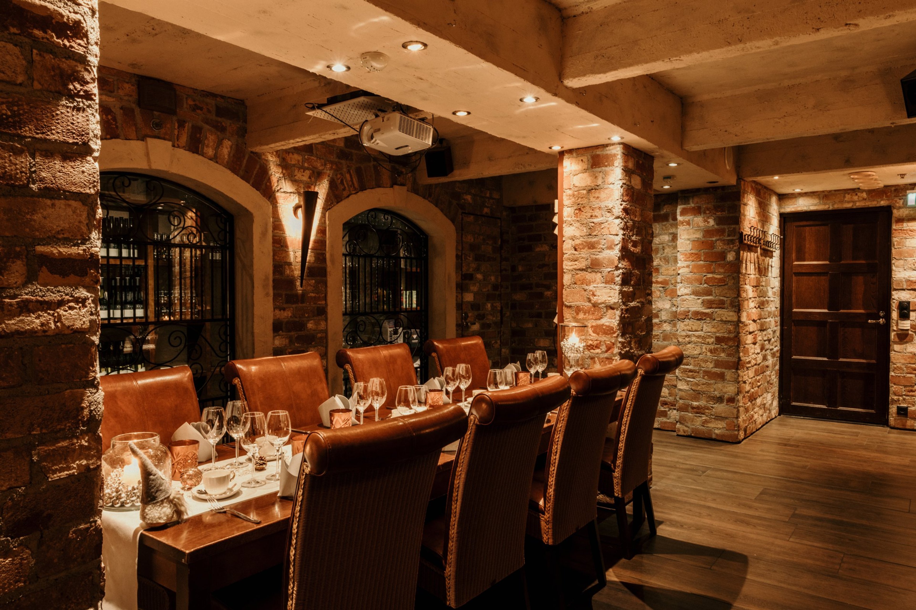 Tunnelmallinen ravintolan yksityishuone tiiliseinillä, kattoprojektorilla ja pitkällä ruokapöydällä nahkatuoleineen. Lattialla puuparketti.