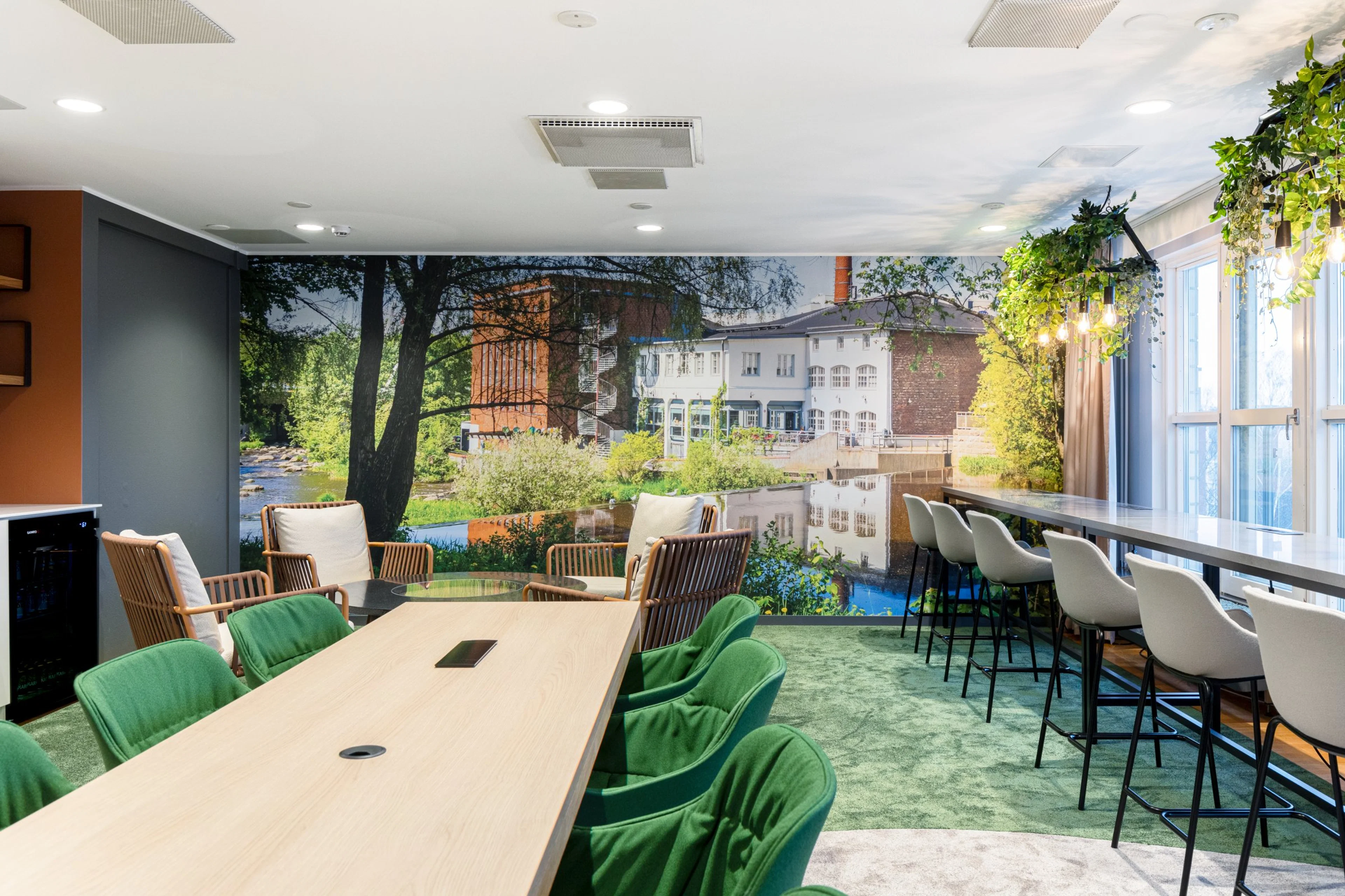 Moderni kokoushuone, jossa puinen pöytä vihreine tuoleineen, korkeita baarituoleja ja seinän kokoinen maisemakuva joesta ja rakennuksista. Vihreä matto ja viherkasvikoristelu.