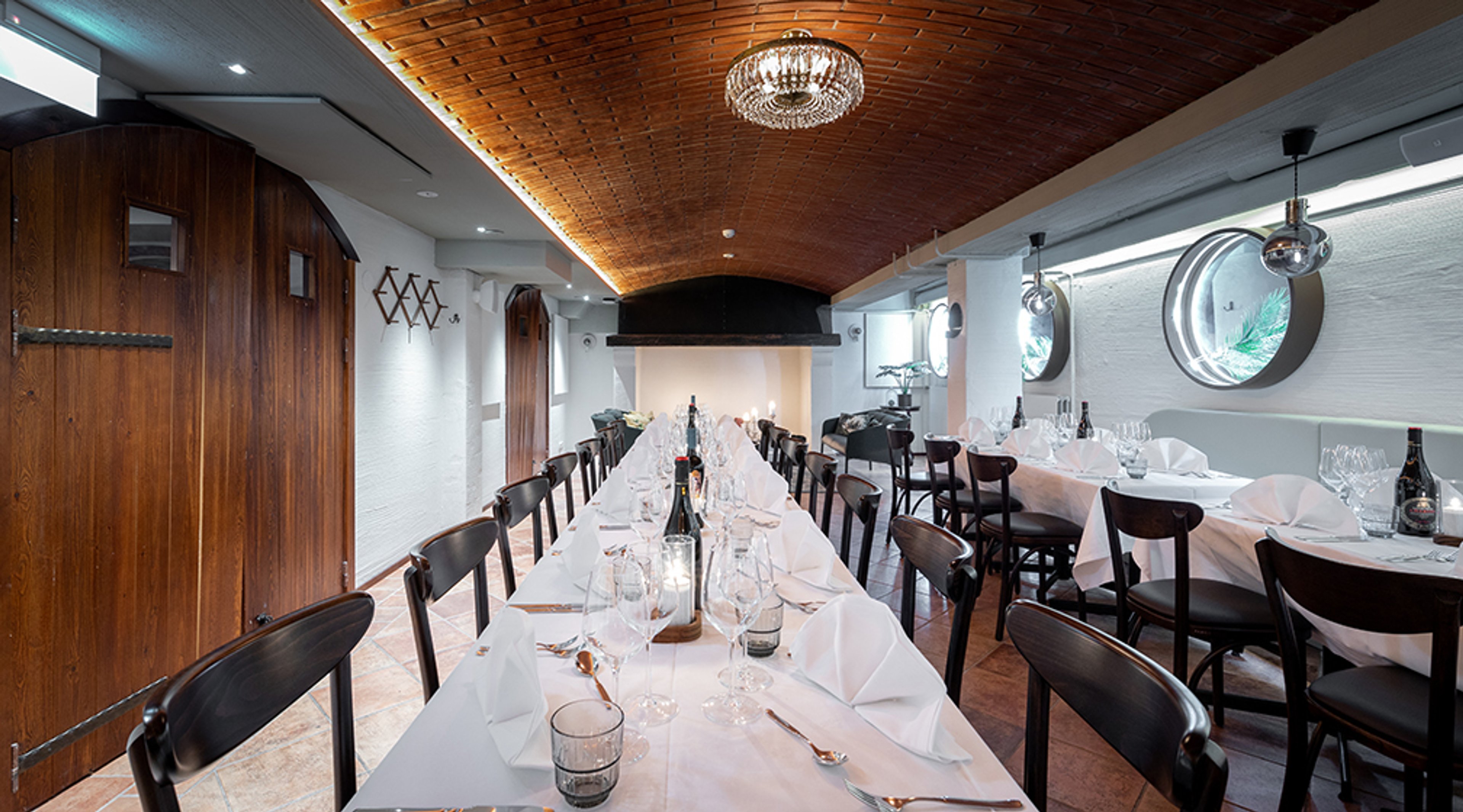 Pitkä pöytä katettu valkoisella liinalla ravintolan yksityistilassa, jossa puinen kaareva katto ja kristallikruunu.