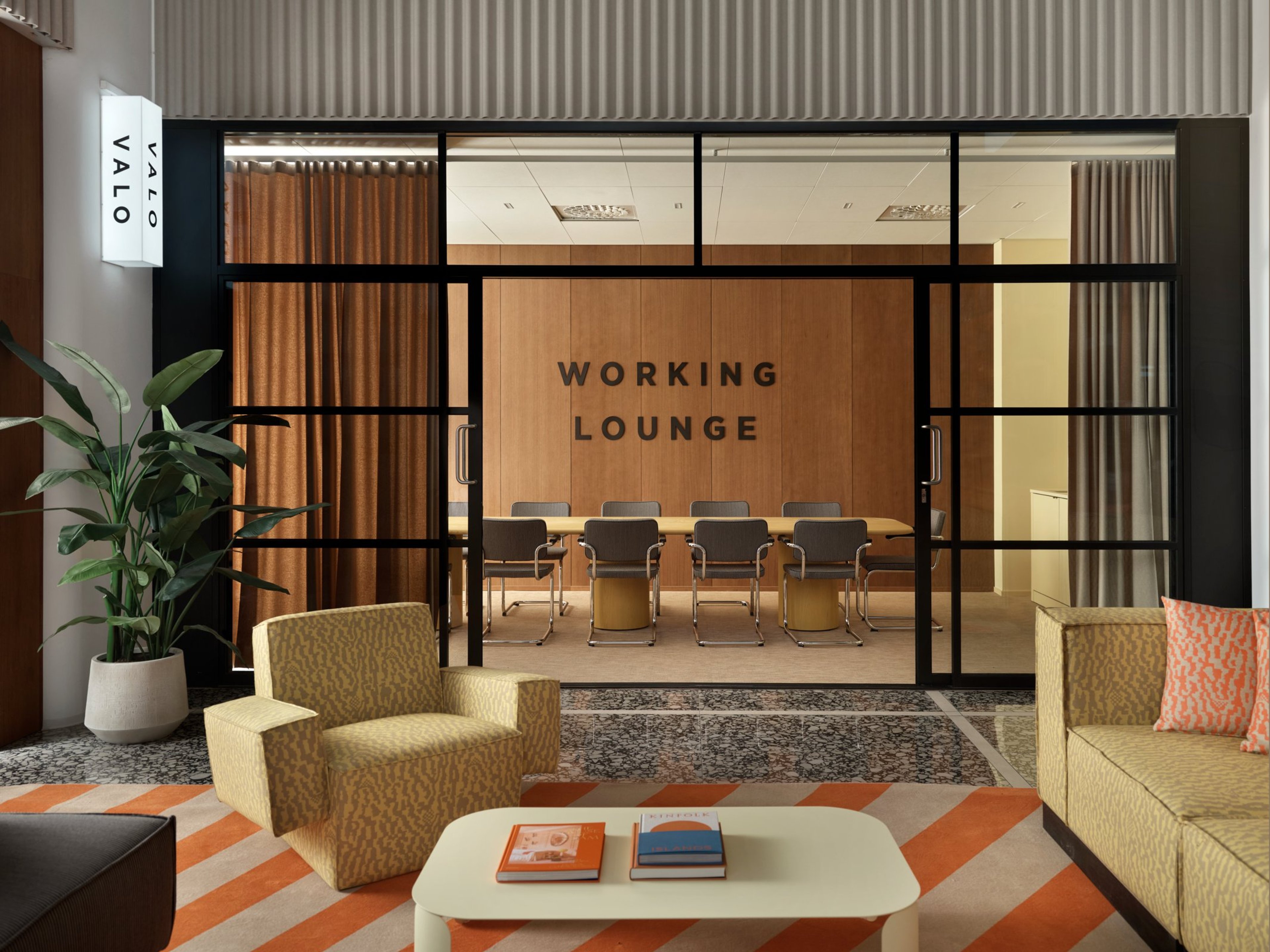 Moderni työskentelytila lasiseinien takana, etualalla oranssi-valkoraidallisen maton päällä oleskelualue sohvineen ja kasveineen.