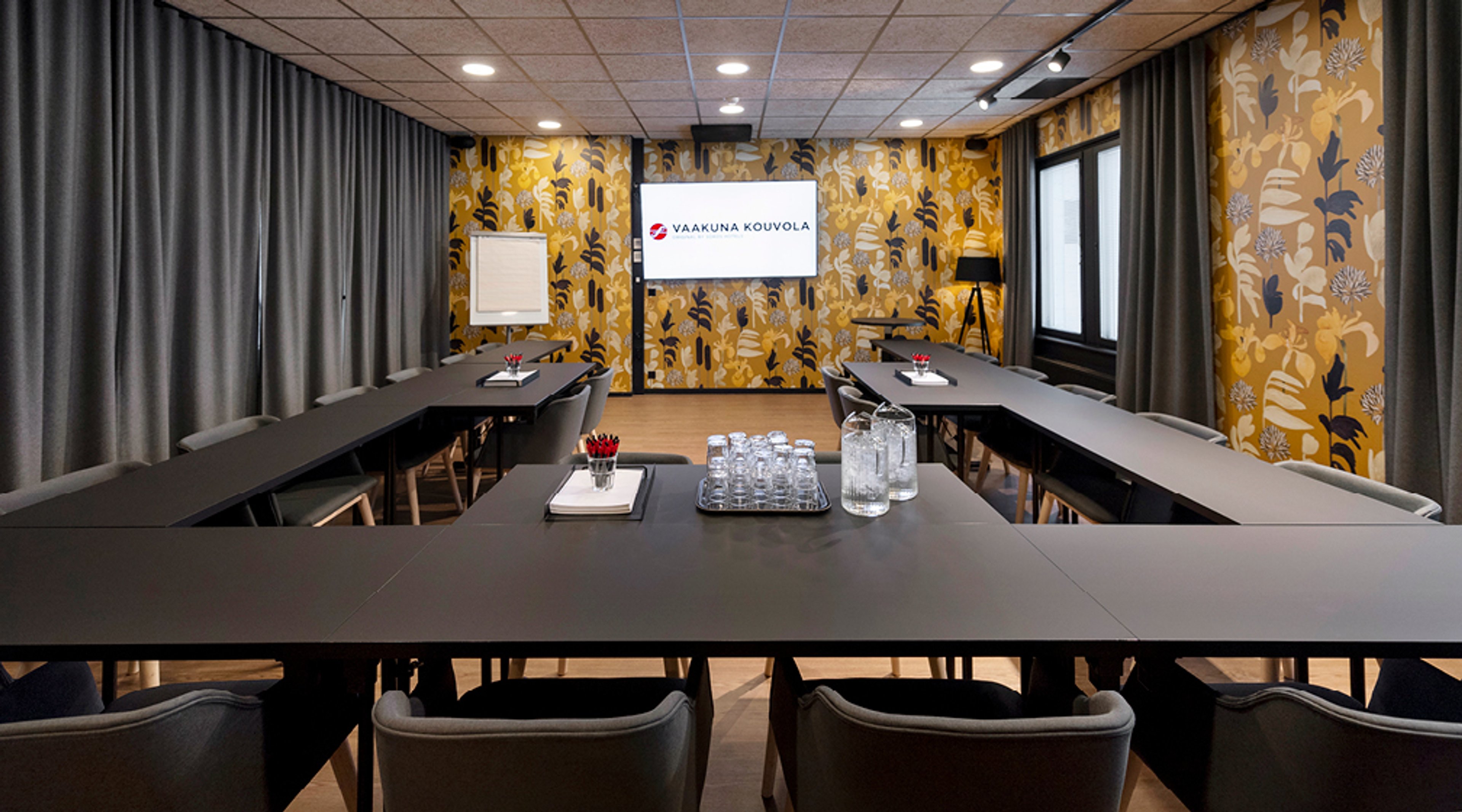 Moderni kokoustila U-muotoisella pöytäasetelmalla, keltaisella kukkakuvioisella seinällä ja harmailla verhoilla. Esillä vettä ja kokoustarpeita.