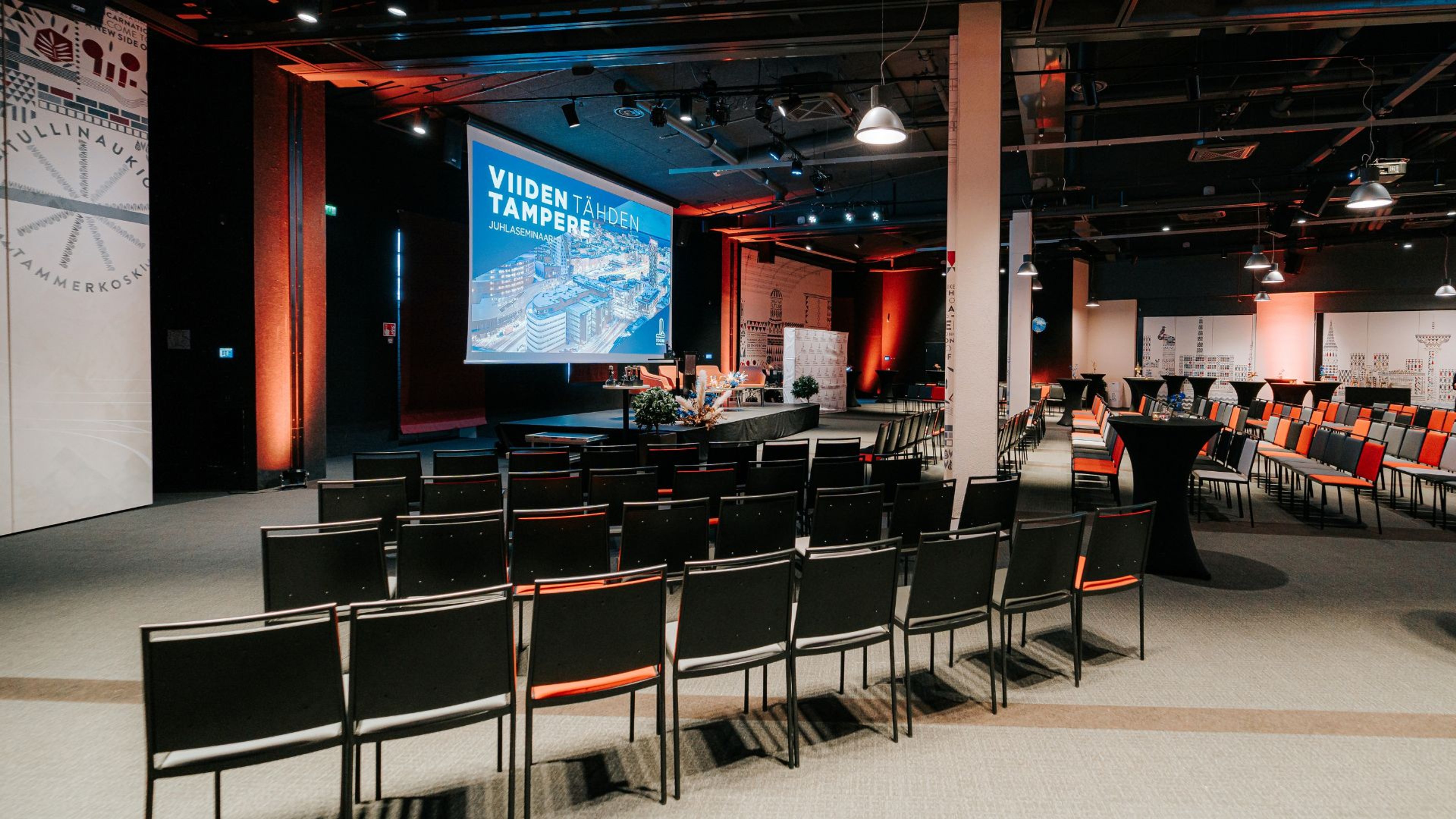 Auditorio tuoleineen ja lavoineen, jossa suuri sininen näyttö. Tila on valaistu punaisella tunnelmavalaistuksella ja lattialla on tasainen kokolattiamatto.