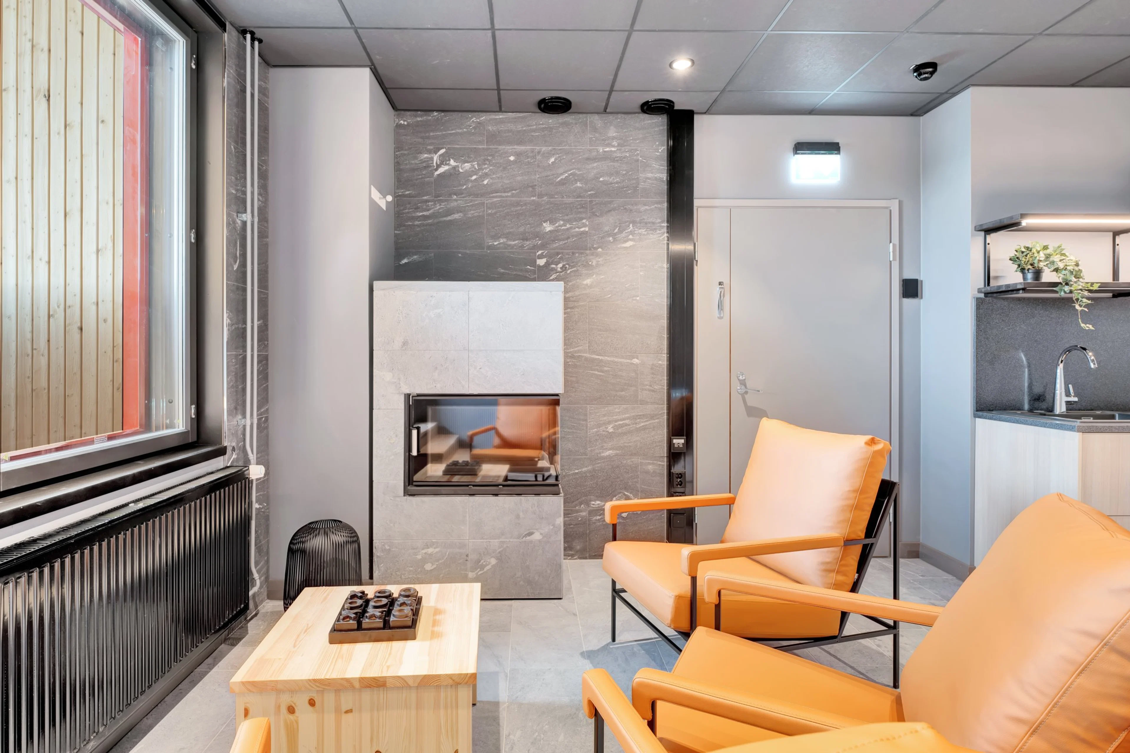 Moderni sauna-alue, jossa marmorikaakeloitu seinä, sisäänrakennettu kiuas, oranssit nojatuolit ja puinen pöytä. Tilassa on myös pieni keittiönurkkaus.