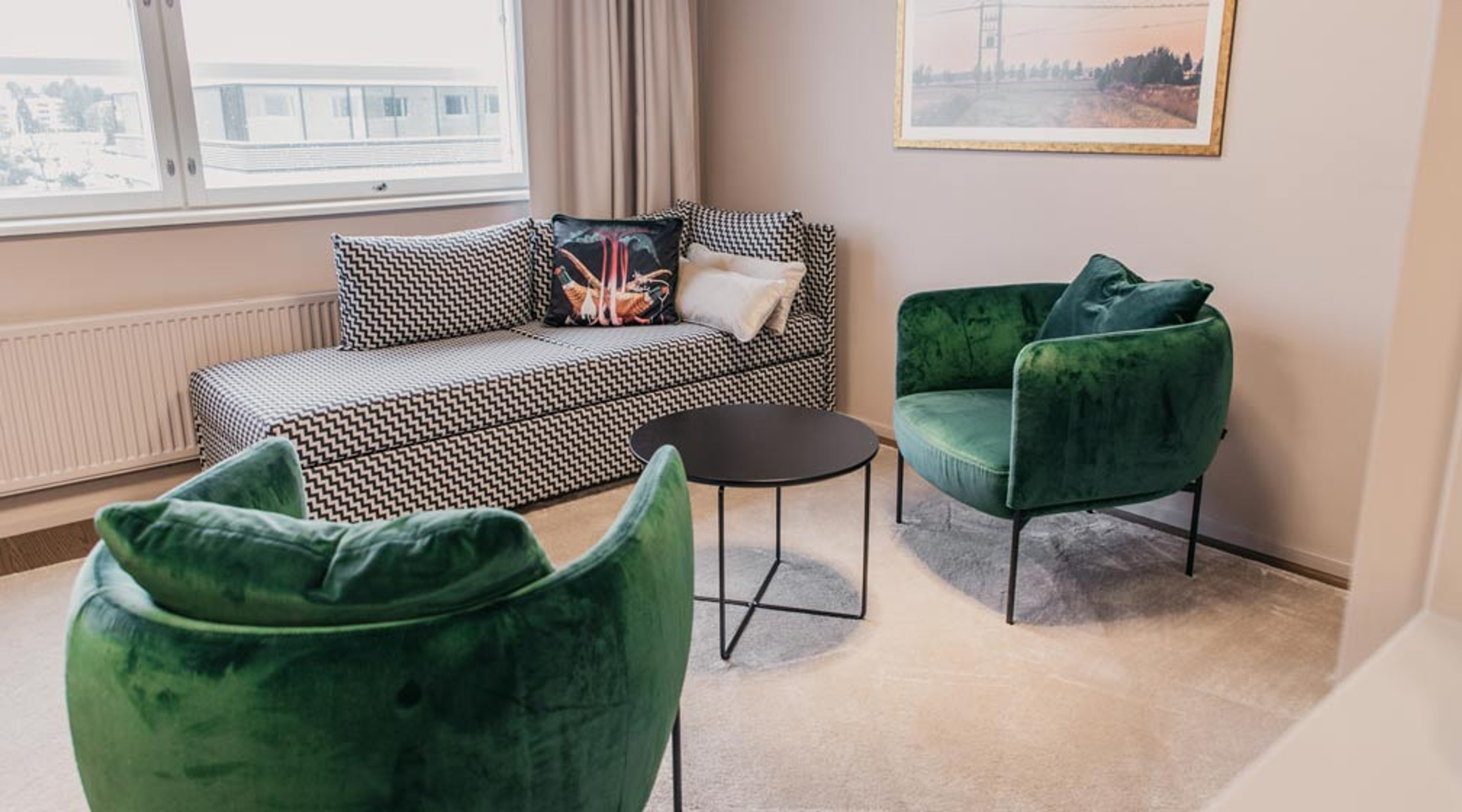 Moderni oleskelutila, jossa kaksi vihreää samettituolia, mustavakoinen sohva, pyöreä sohvapöytä ja ikkunanäkymä. Vaalea lattia ja ohut matto.