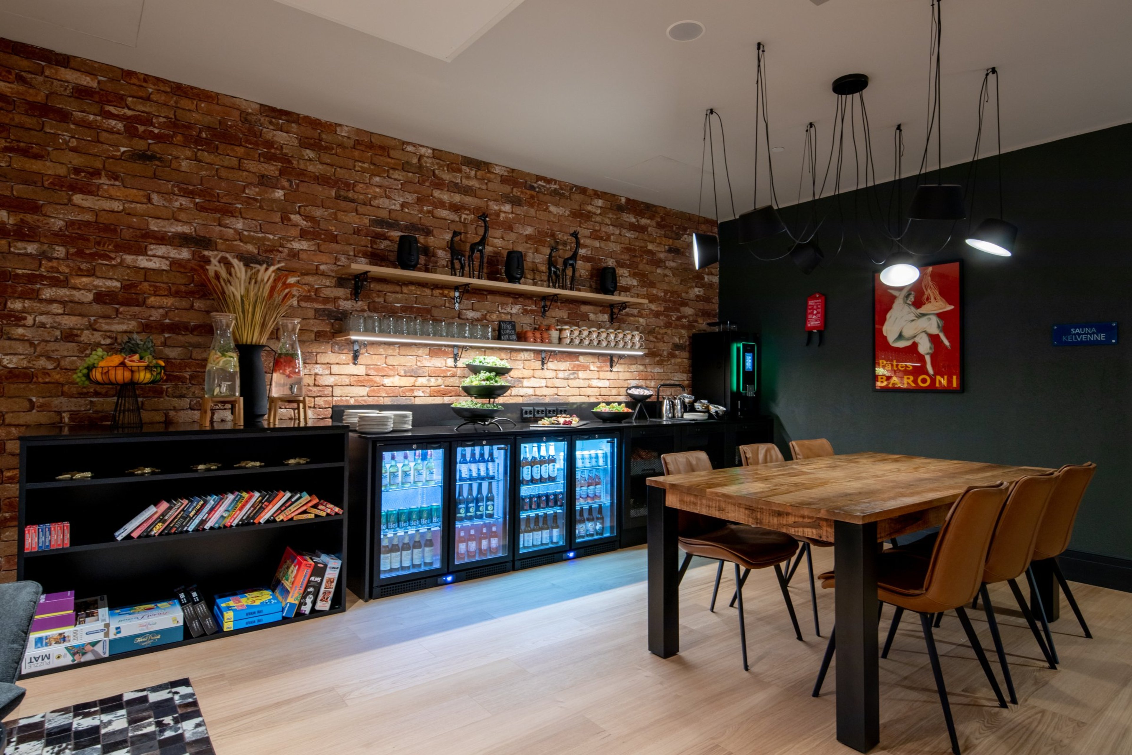 Tunnelmallinen tila tiiliseinällä, juomakaapit, puupöytä nahkatuoleineen, avoin lattiatila ja vintage-julisteet seinällä.