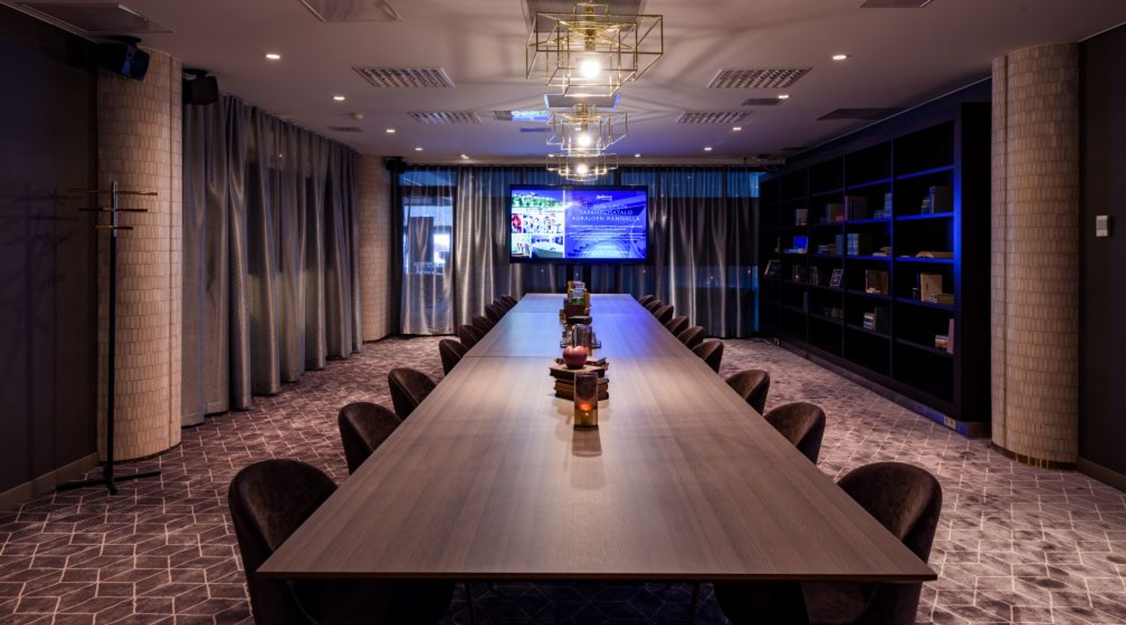 Moderni neuvotteluhuone pitkällä puupöydällä ja tummanruskeilla tuoleilla. Taustalla verhojen edessä näyttö ja sivulla kirjahylly. Tilassa hyvä valaistus ja tasainen lattia.
