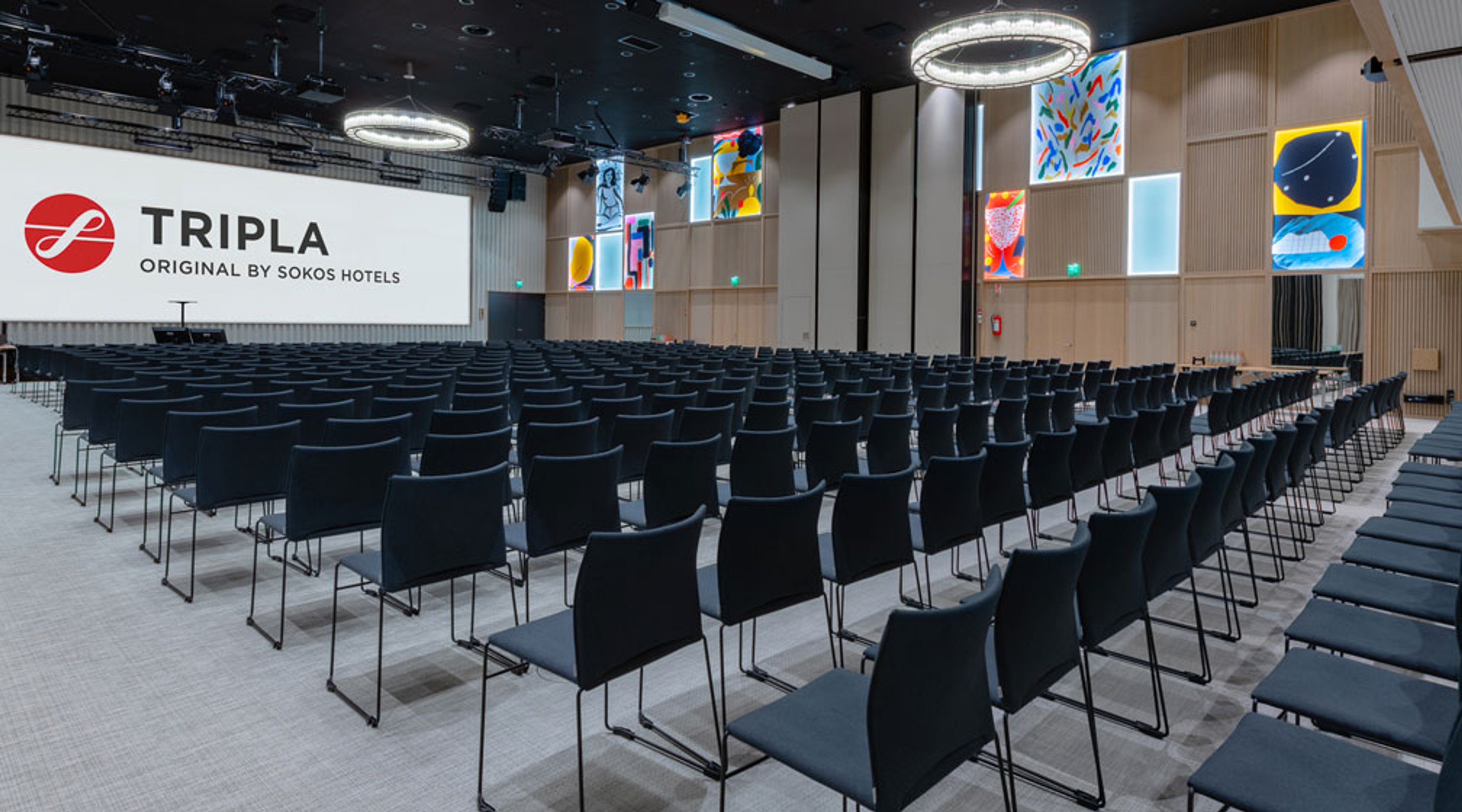 Moderni auditorio mustilla tuoleilla, puupaneeleilla, taideteoksilla seinillä ja valaisevilla rengaskruunuilla. Tasainen lattia helpottaa liikkumista.