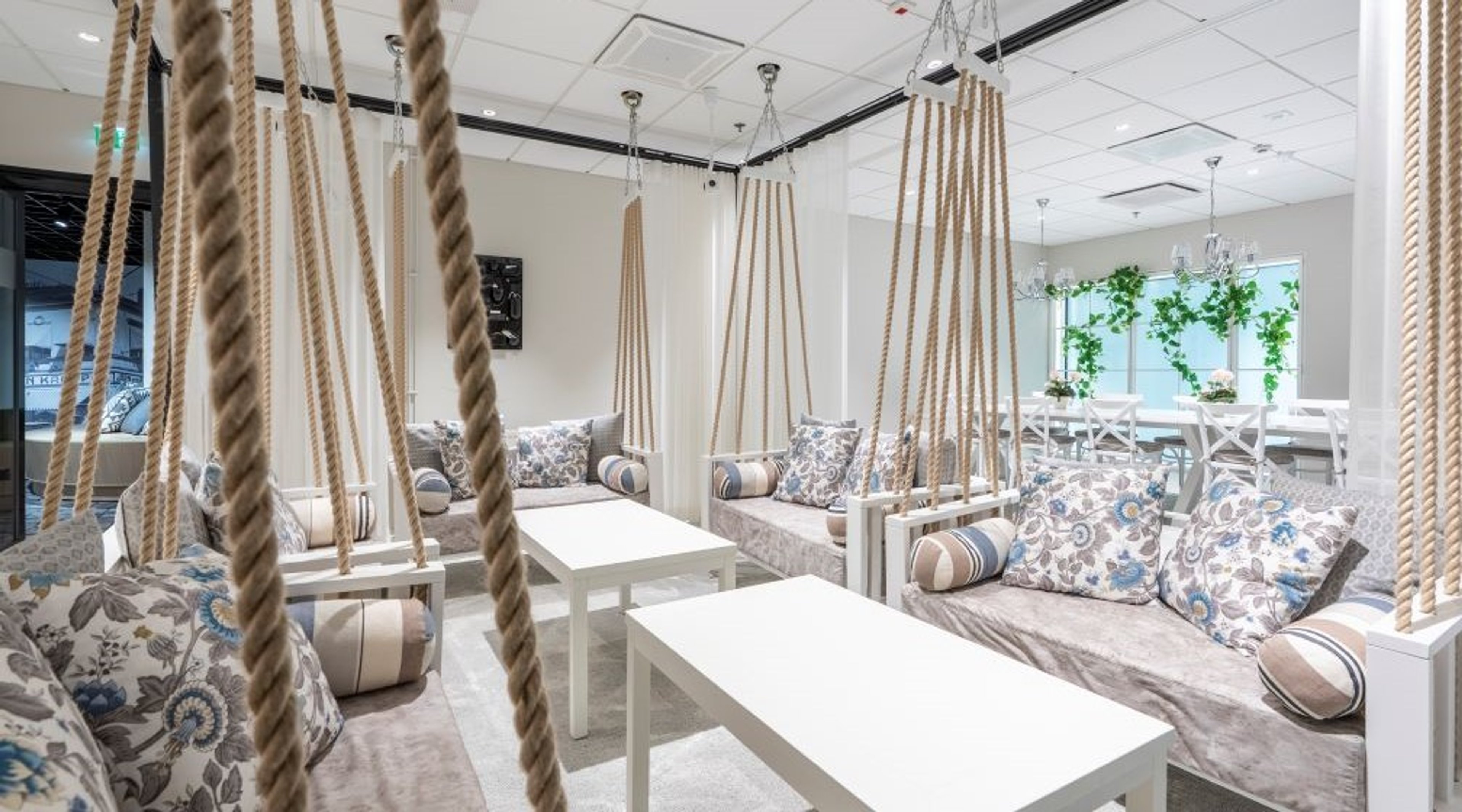 Lounge-tila, jossa katosta roikkuvia köysillä ripustettuja sohvia kukallisilla tyynyillä ja valkoisilla pöydillä. Ikkunoita ja viherkasveja taustalla.