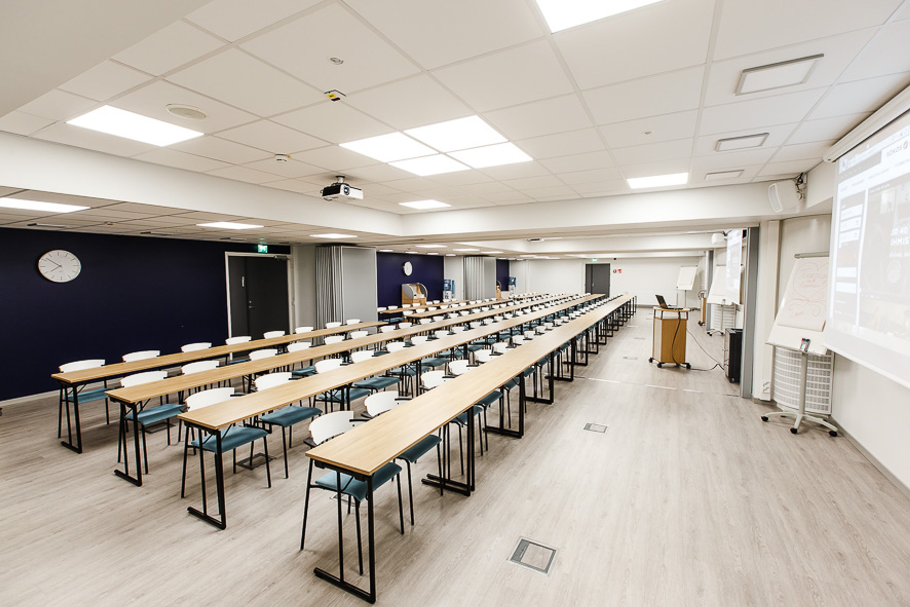 Valoisa luentosali, jossa on pitkät pöydät tuoleineen riveittäin, projektori katossa ja esitysvalkokangas oikealla. Tilassa on puulattia ja tummansininen takaseinä.