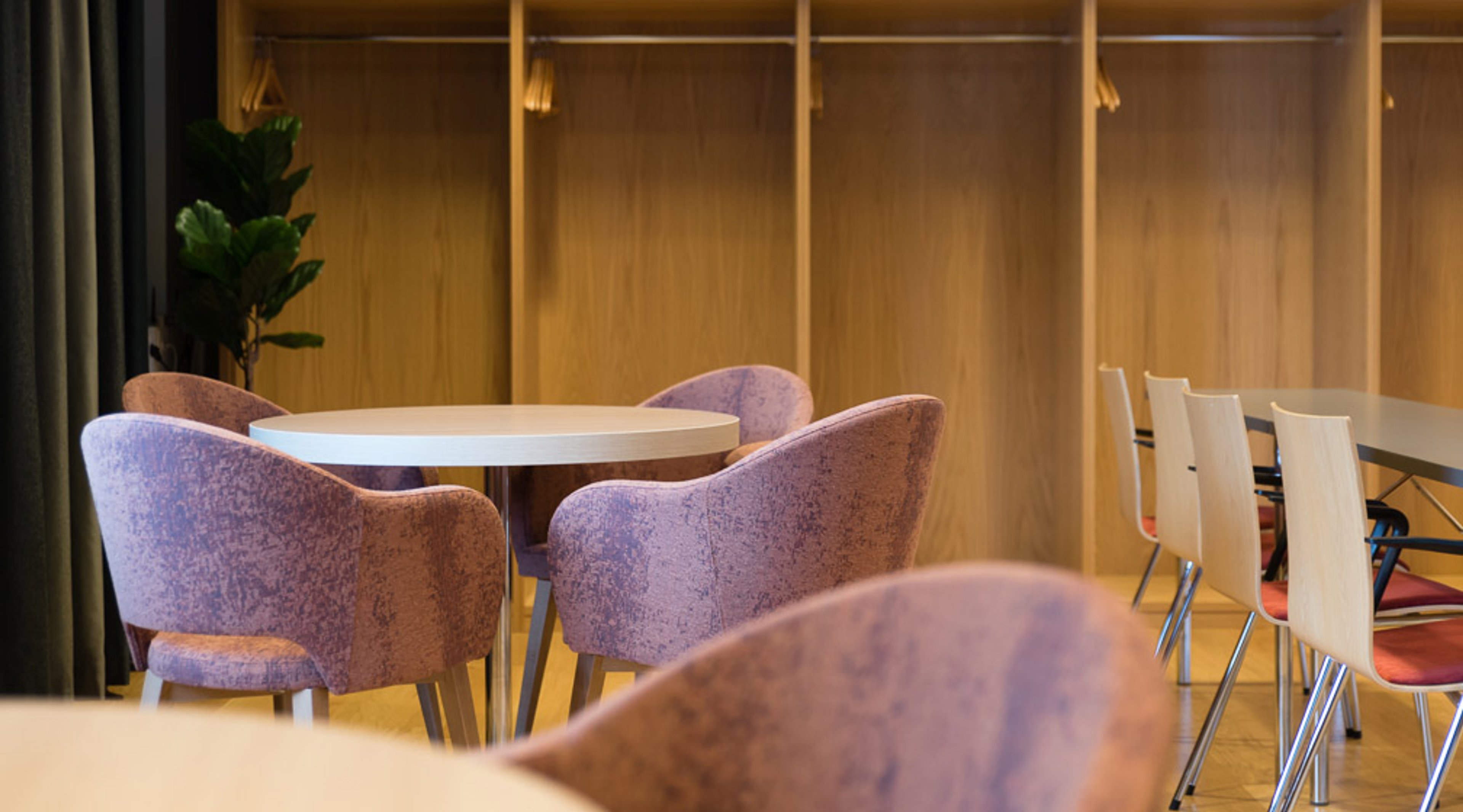 Moderni kokoustila pyöreällä pöydällä ja violeteilla nojatuoleilla, taustalla puinen seinärakenne ja viherkasvi.