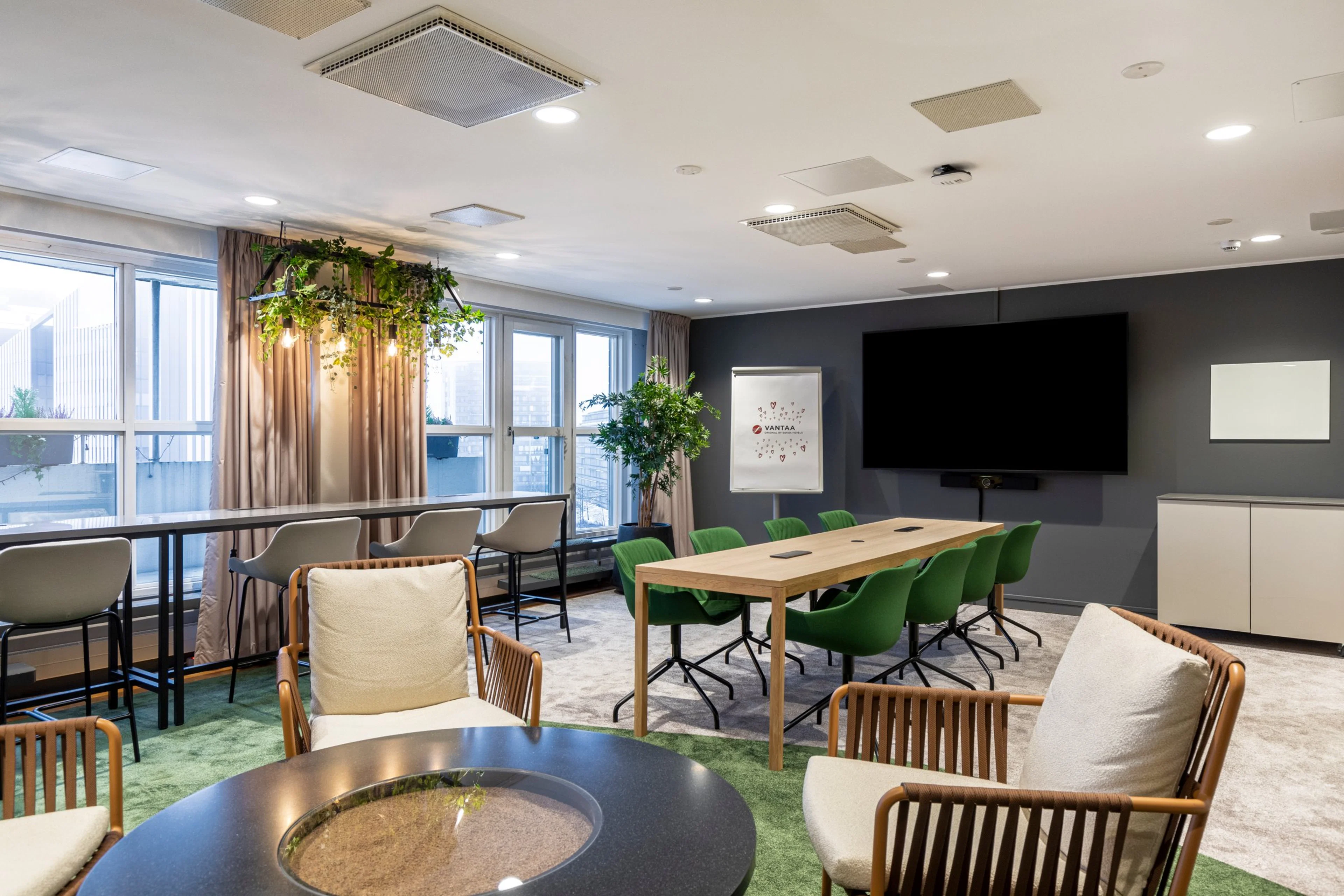 Moderni neuvotteluhuone, jossa puupöytä vihreillä tuoleilla, mukavia nojatuoleja, kasvikattovalaisin ja iso näyttö tummalla seinällä.