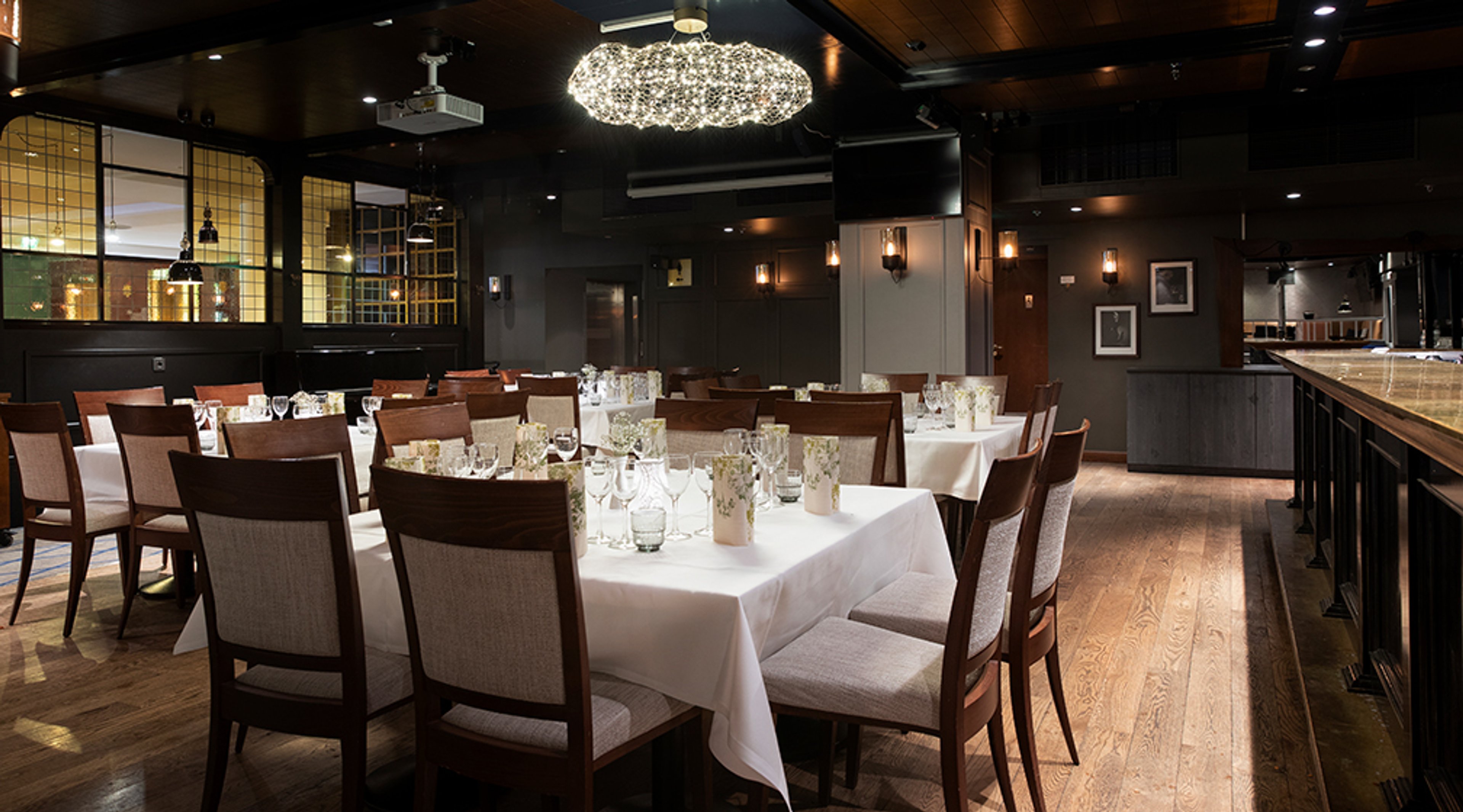 Tyylikäs ravintolasali, jossa puulattia, tummat seinät ja kattaus valkoisilla pöytäliinoilla. Kattokruunu valaisee tilaa.