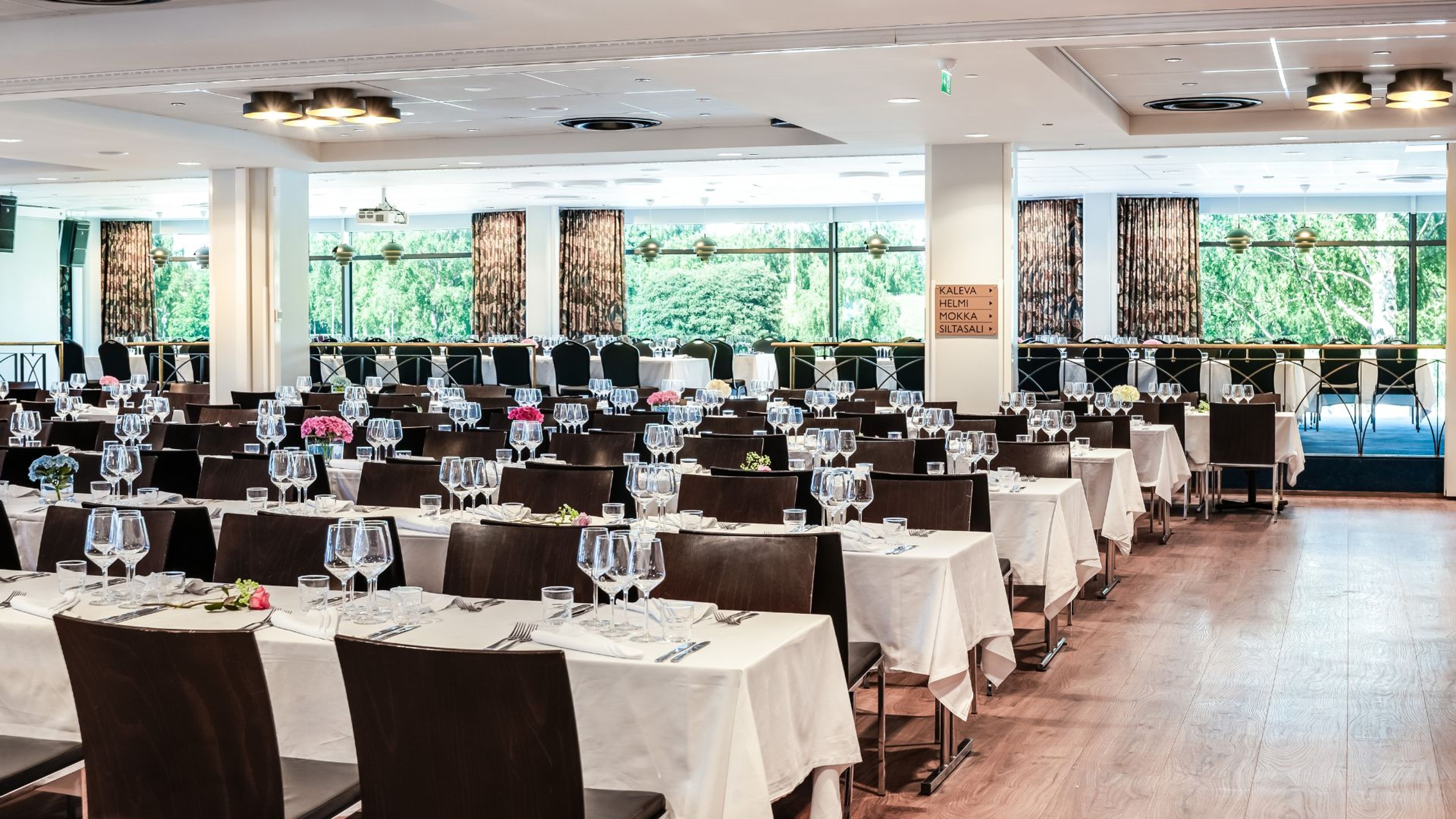 Valoisa ravintolasali tummin tuolein ja valkoisin pöytäliinoin. Pöydillä viinilaseja ja pieniä kukkia. Suurista ikkunoista näkyy vehreä maisema.