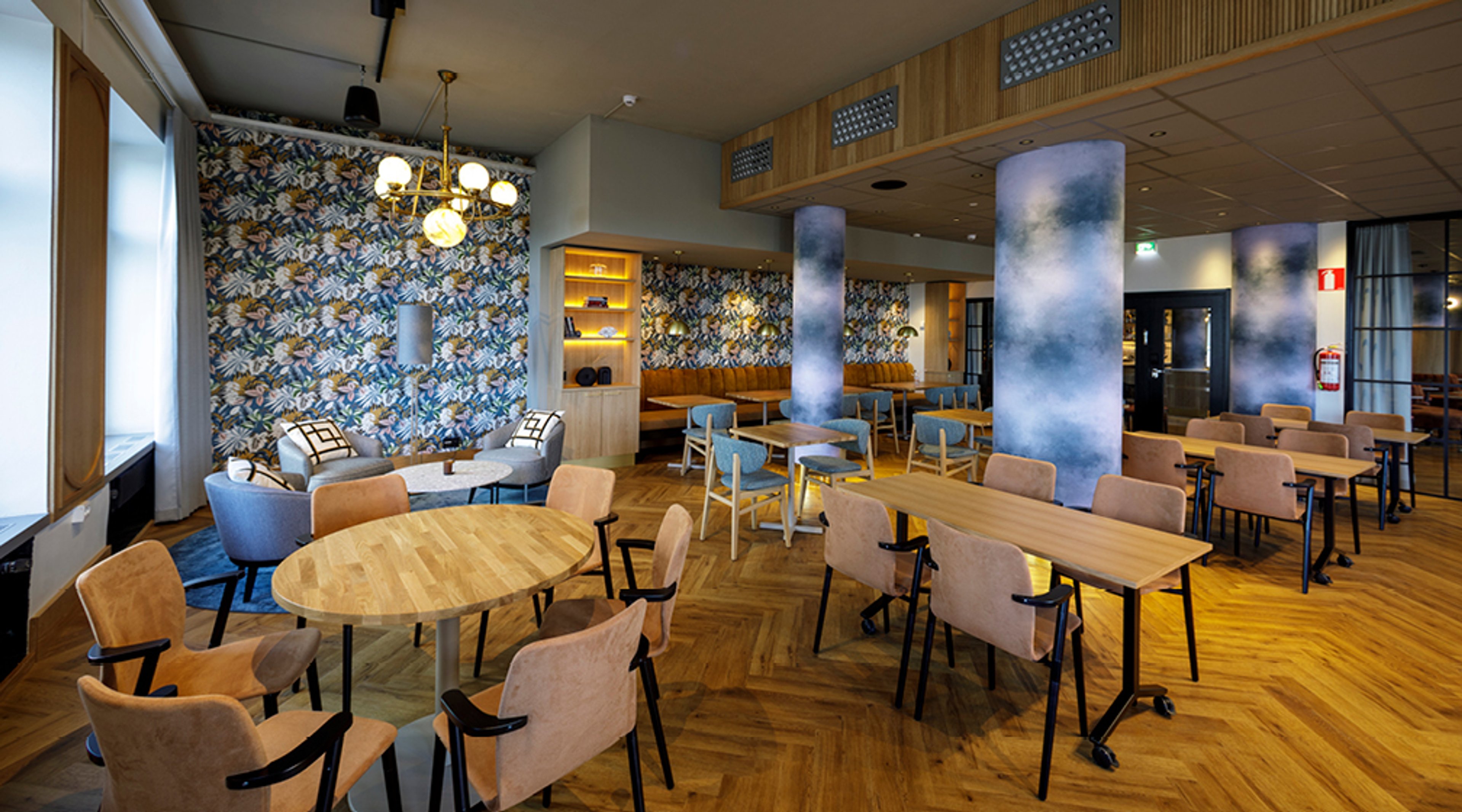 Moderni kahvila puulattialla. Seinillä kukkakuvioinen tapetti, keskellä pyöreät pöydät ja tuolit. Tila on valoisa ja avara.