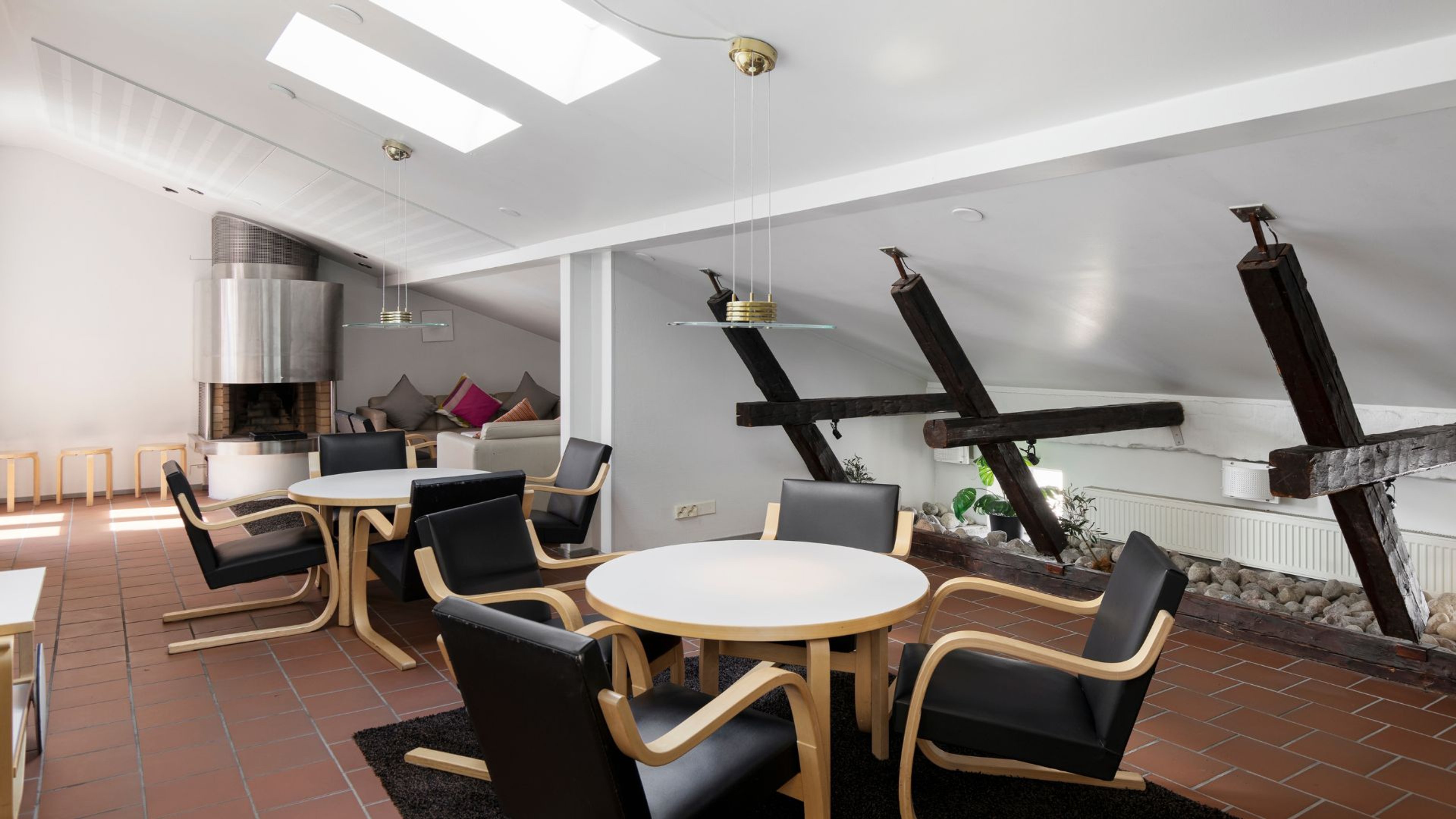 Moderni loungealue, jossa pyöreät pöydät, mustat nahkanojatuolit, tiiliseinä, kattovalot ja takka. Tilassa näkyy puiset kattopalkit ja kattoikkunat.