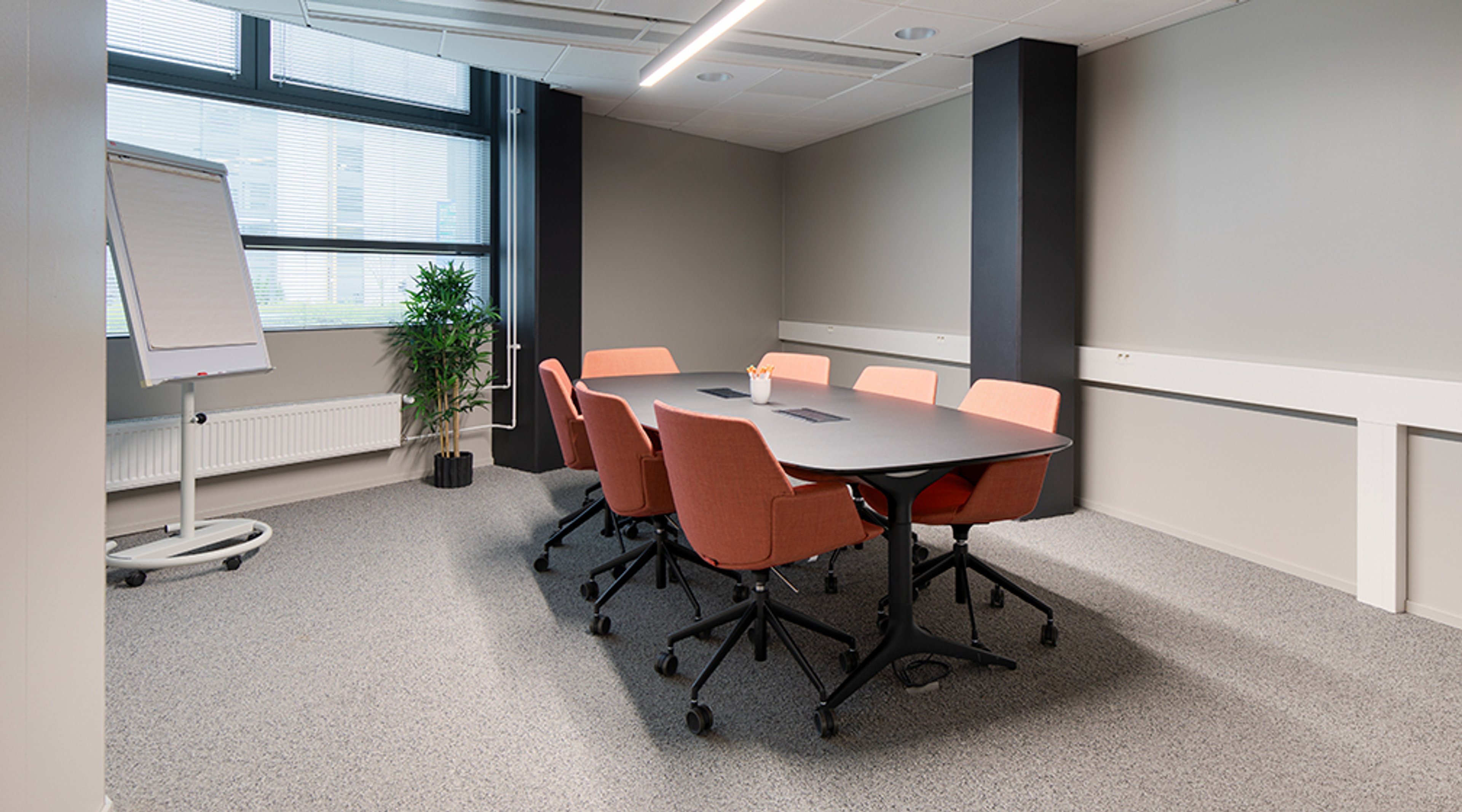 Moderni neuvotteluhuone, jossa on oranssinväriset tuolit ovaalin pöydän ympärillä, fläppitaulu ja viherkasvi ikkunan vieressä. Huoneessa on harmaa lattiamatto ja hyvä valaistus.