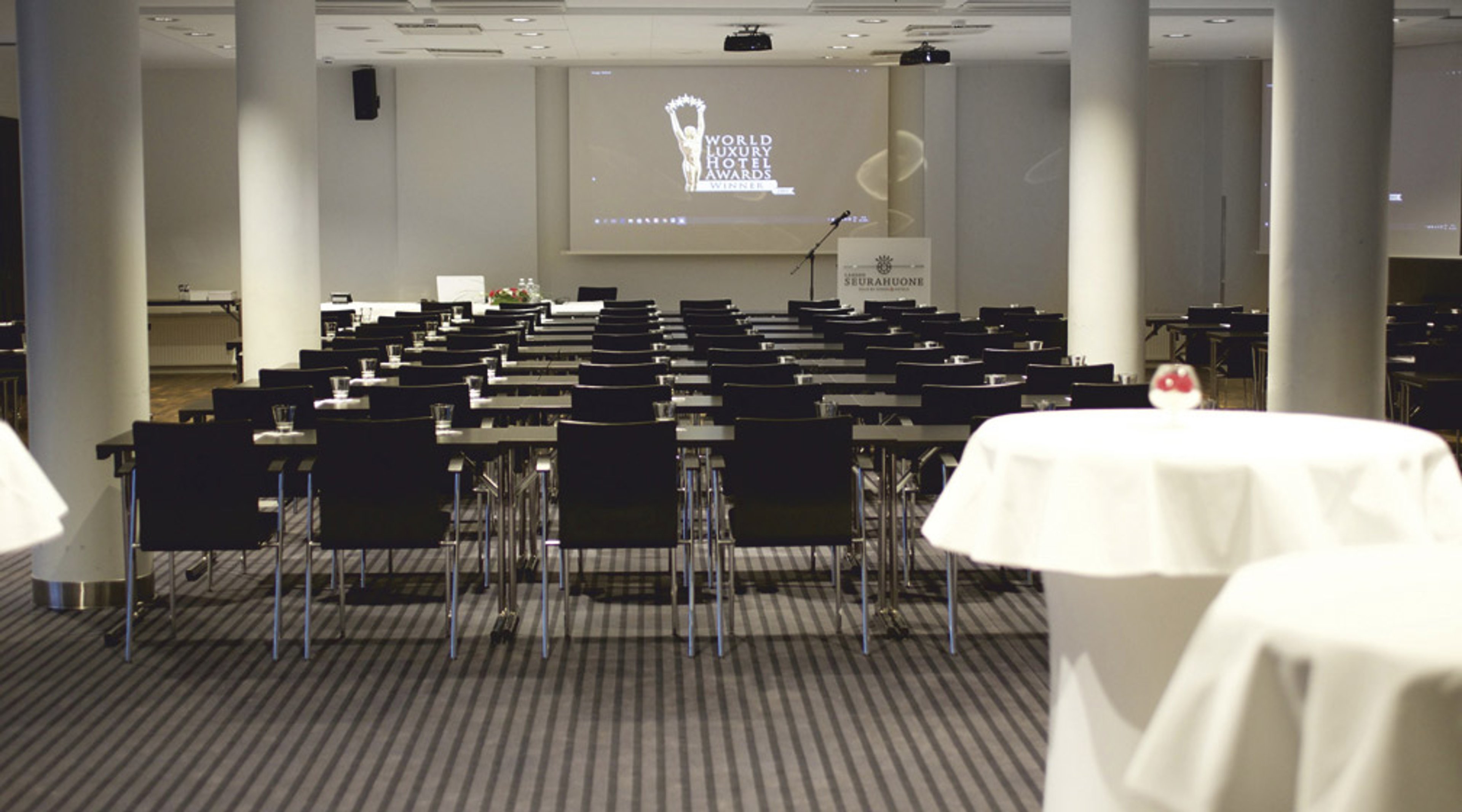 Konferenssihuone, jossa on rivejä mustia tuoleja, valkoisia pöytiä ja valkokangas projektorilla. Kaksi valkoista cocktailpöytää etualalla.