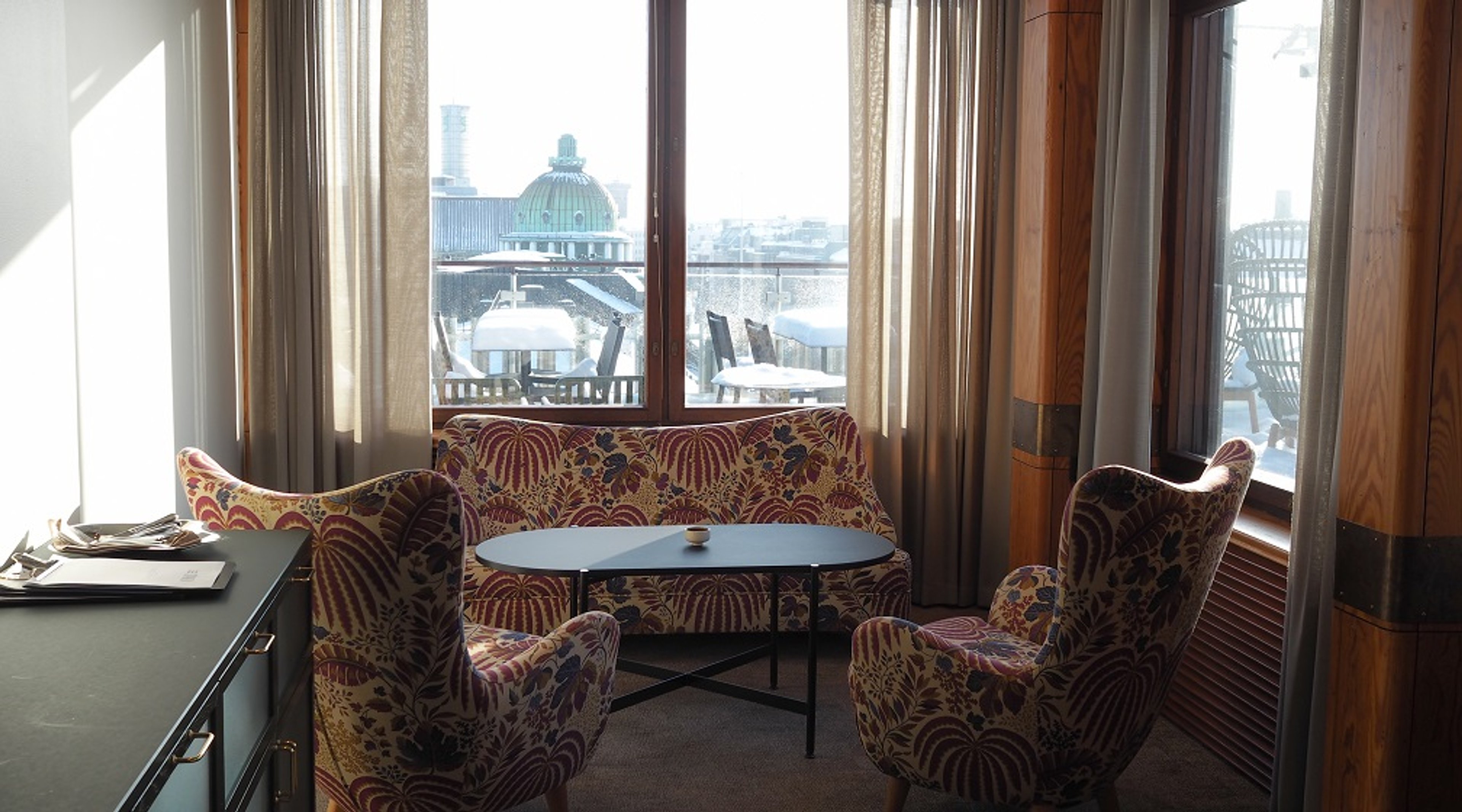 Hotellihuoneen oleskelutila värikkäillä kukkakuvioisilla kalusteilla. Ikkunasta näkyy vihreäkupolinen rakennus.