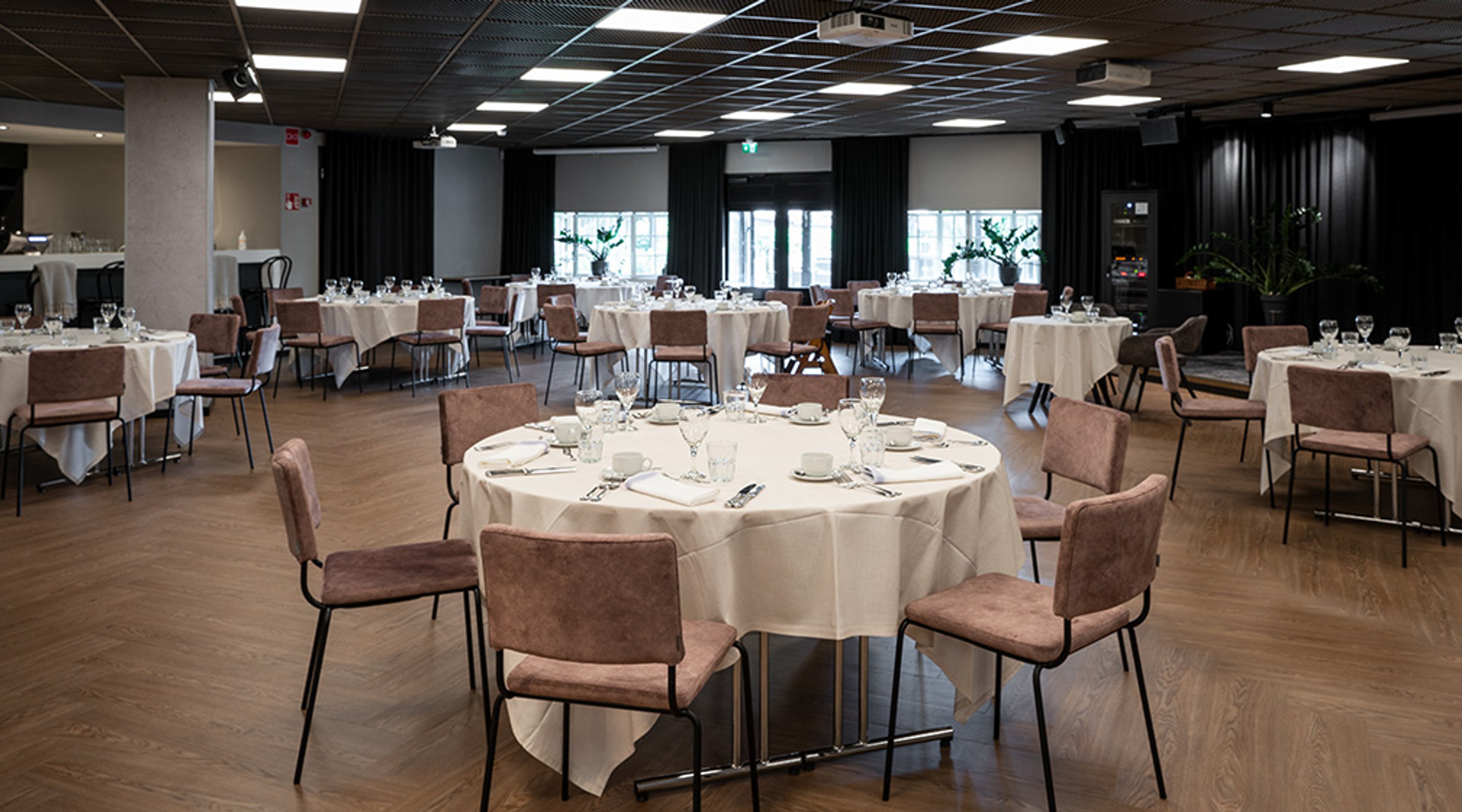 Tilava juhla- tai konferenssisali, jossa useita pyöreitä pöytiä valkoisilla pöytäliinoilla ja ruskeilla tuoleilla. Puulattia ja ikkunat taustalla.