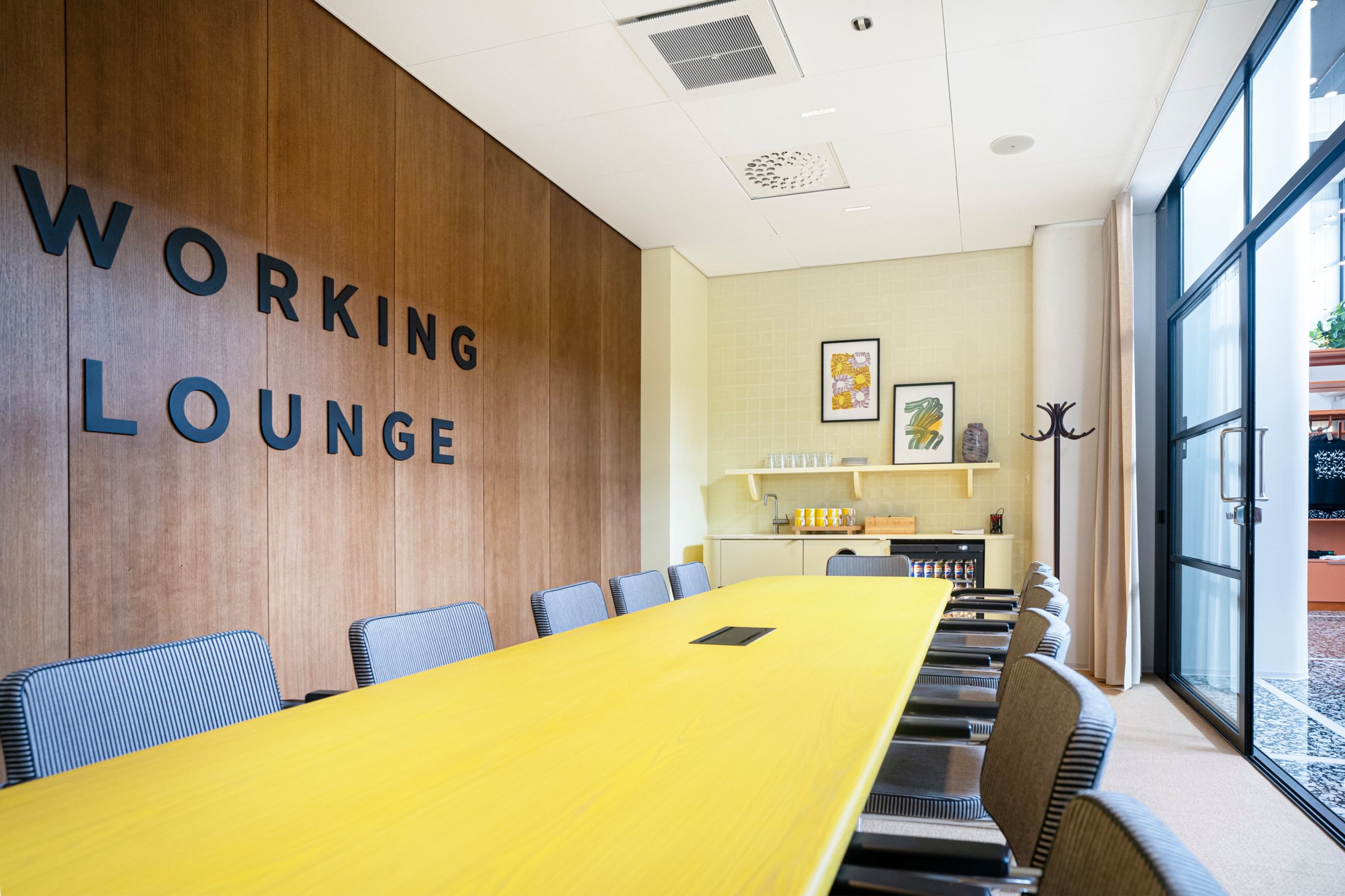 Moderni yhteistyötila, jossa on keltainen neuvottelupöytä, raidoitettuja tuoleja, puupaneeliseinä, keittiönurkkaus ja isot ikkunat. Tasainen lattia ja leveät kulkutilat.