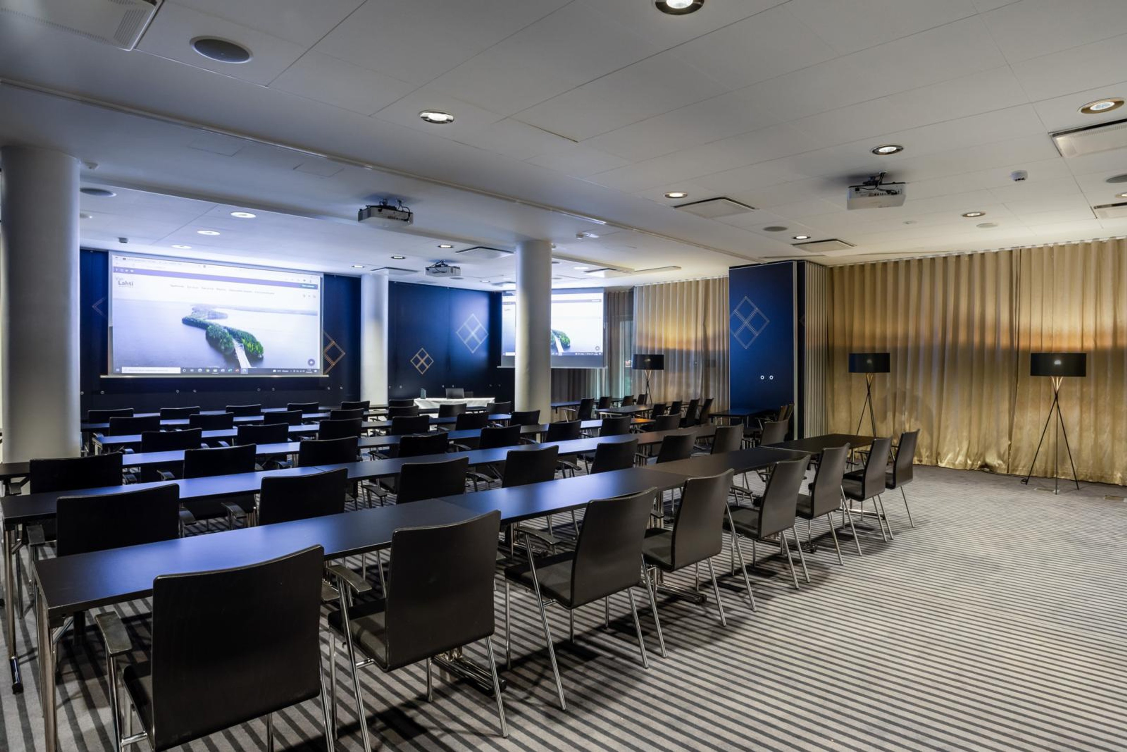 Moderni kokoustila, jossa pitkät pöydät ja tuolit rivissä, sininen taustaseinä, projektoreita ja valkokangas. Tilassa hyvä valaistus ja tasainen lattia.