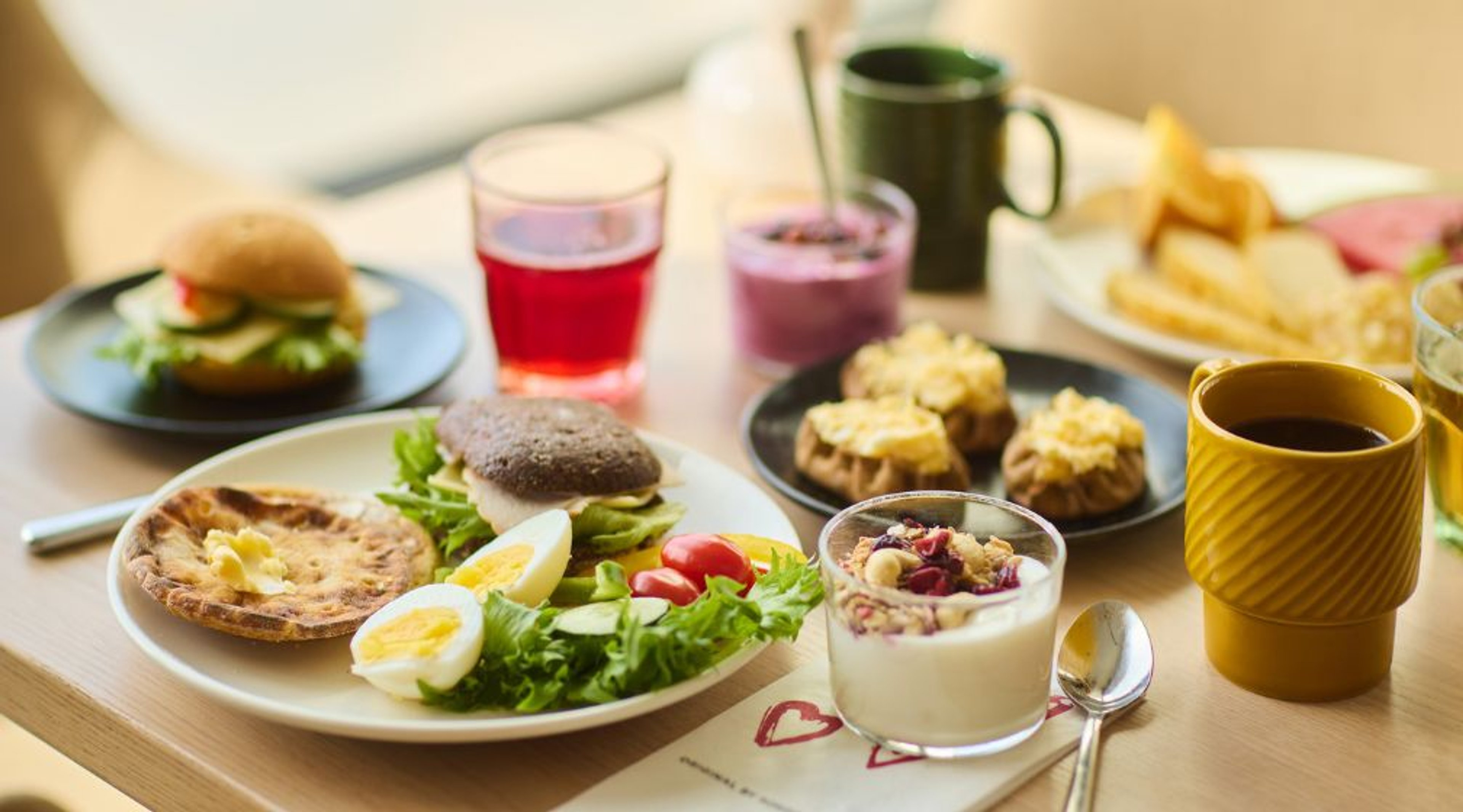 Ett mångsidigt frukostbord med hamburgare, fyllda svampar, sallad, smoothie, kaffe och andra drycker på ett ljust träbord.