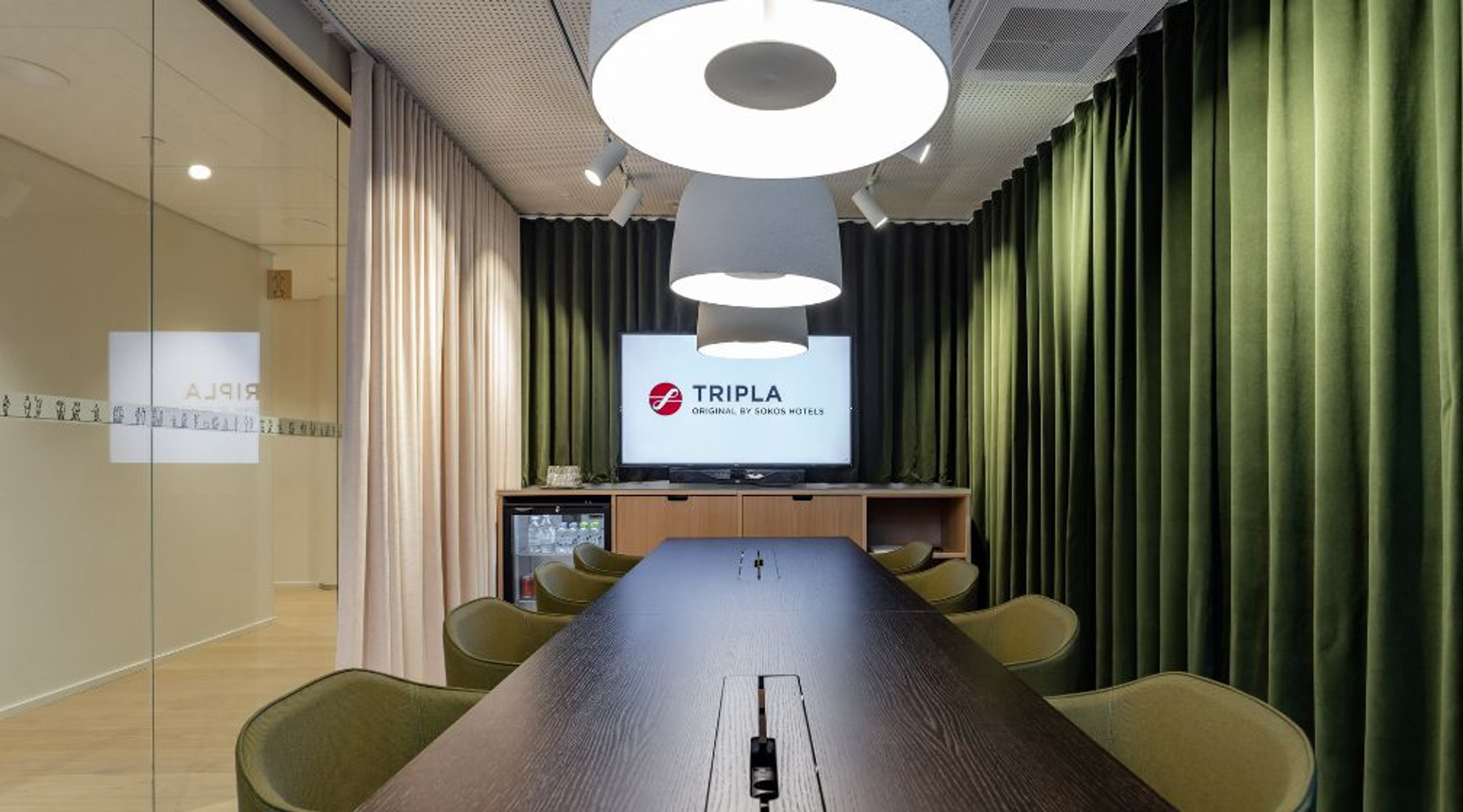 Moderni kokoushuone, jossa tumma puinen pöytä, vihreät tuolit ja verhot. Kattolamppu roikkuu pöydän yläpuolella. Lasiseinä vasemmalla.