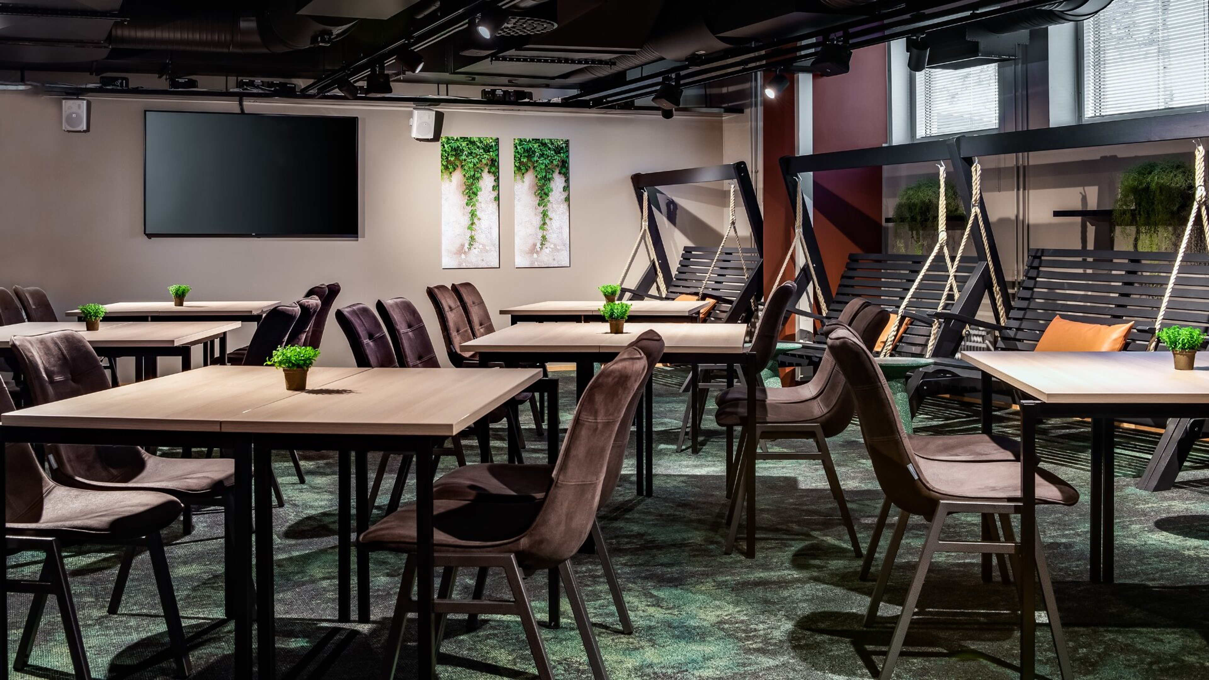 Moderni kokoustila pöytäryhmillä, näytöllä ja keinuistuimilla. Vihreitä kasveja pöydillä ja tauluja seinillä. Tasainen lattia ja runsaasti tilaa liikkua.
