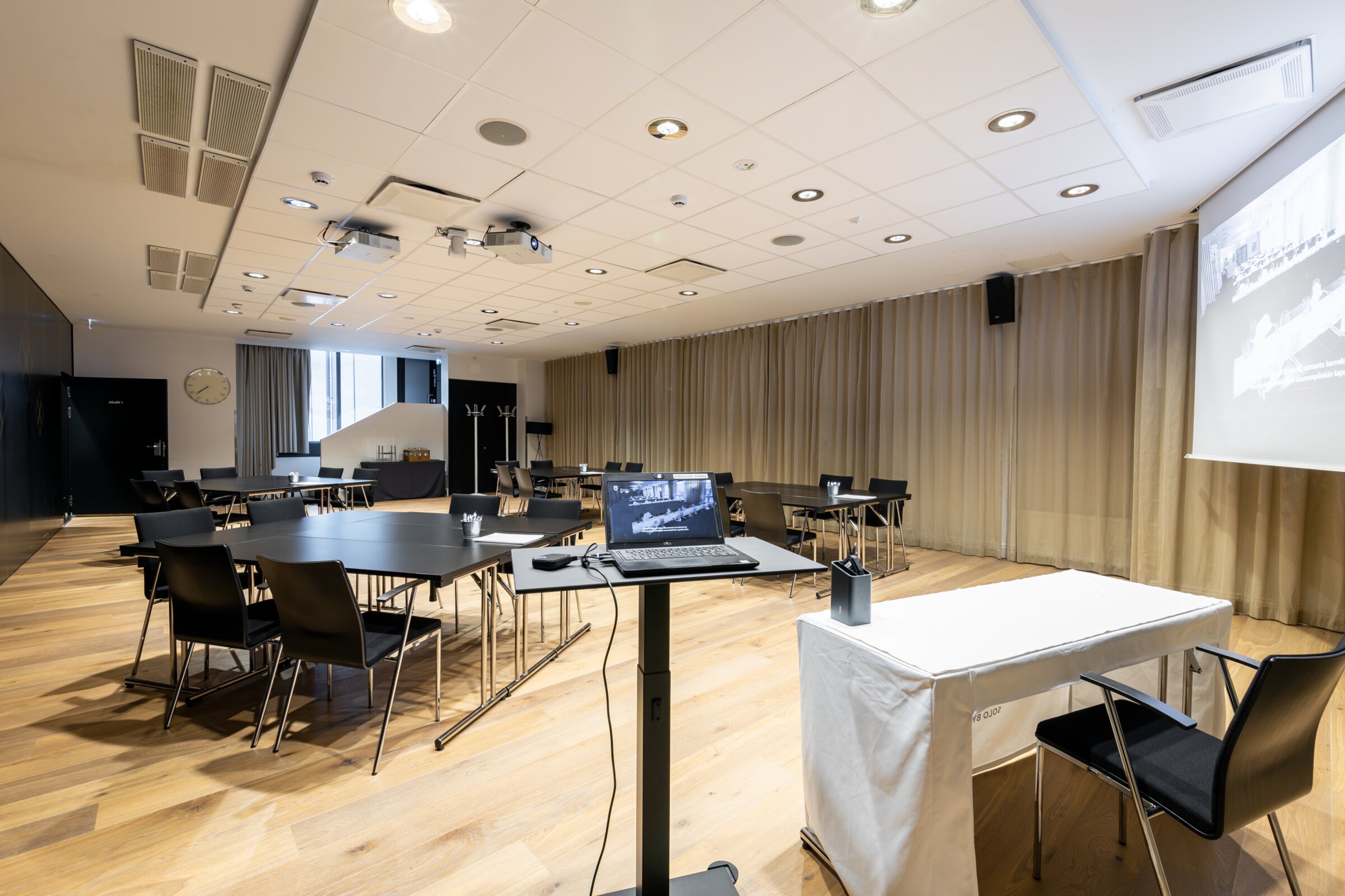 Moderni kokoustila puulattialla, jossa pöytiä ja tuoleja. Tilassa valkokangas, projektori katossa ja läppäri esityspöydällä.